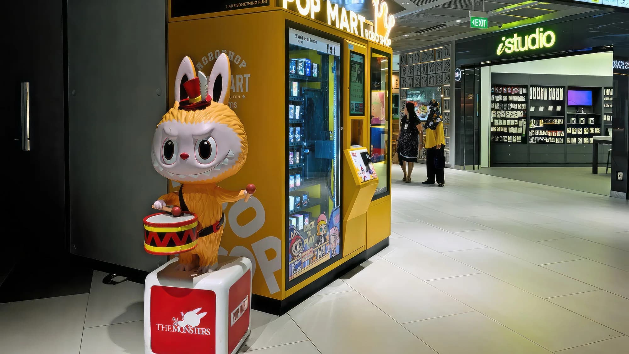 A brand activation in a shopping mall: a Pop Mart Monsters figure with a drum stands beside a yellow vending shop.