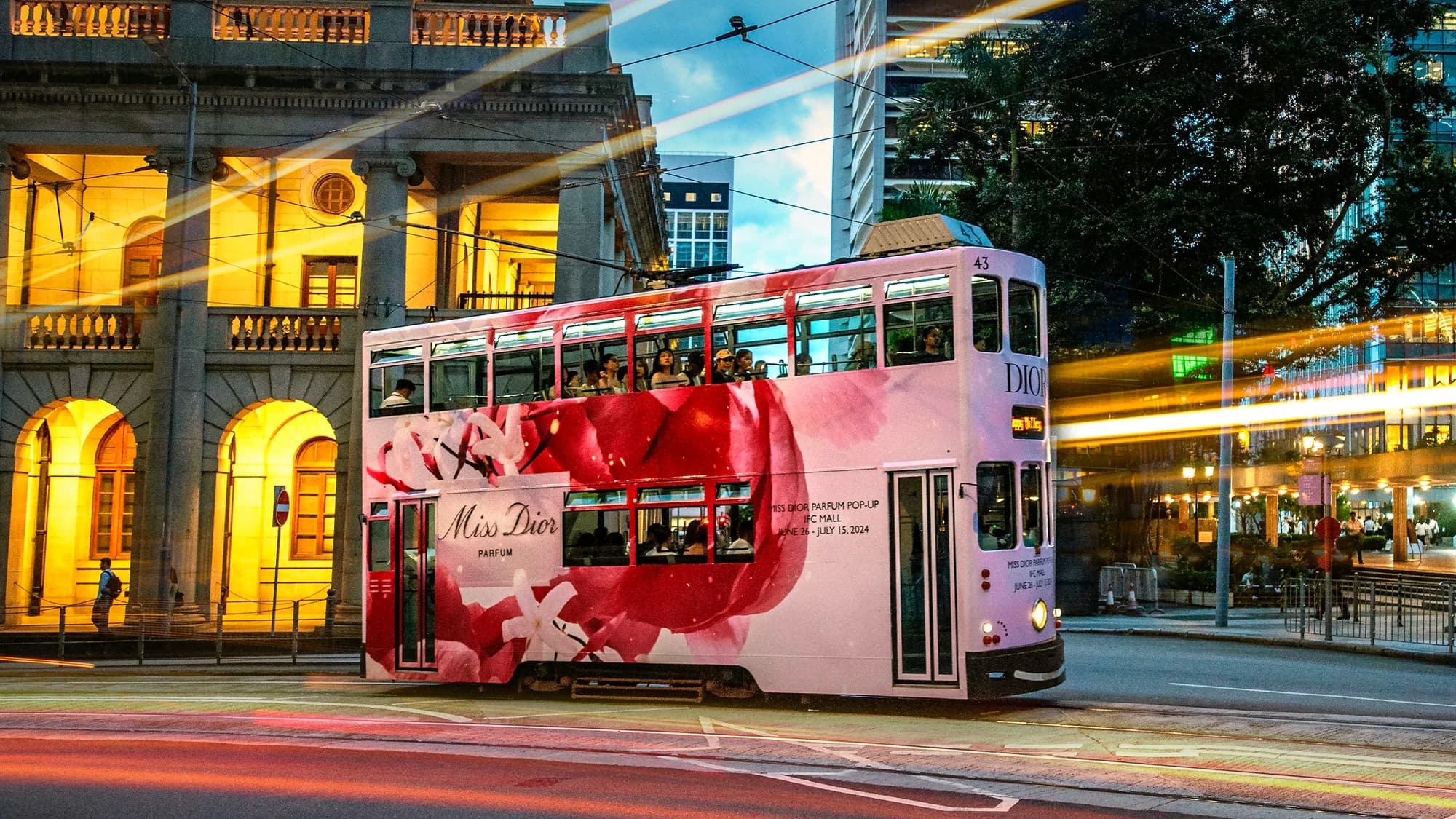 Tram advertising: a double-decker tram wrapped in pink Miss Dior advertising runs past a lit stone building.