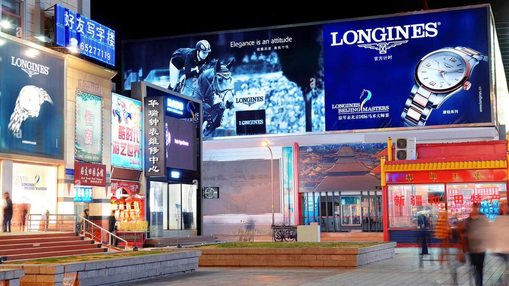 Billboard advertising in Wangfujing, Beijing: Longines boards reading elegance is an attitude cover a night-time building front.