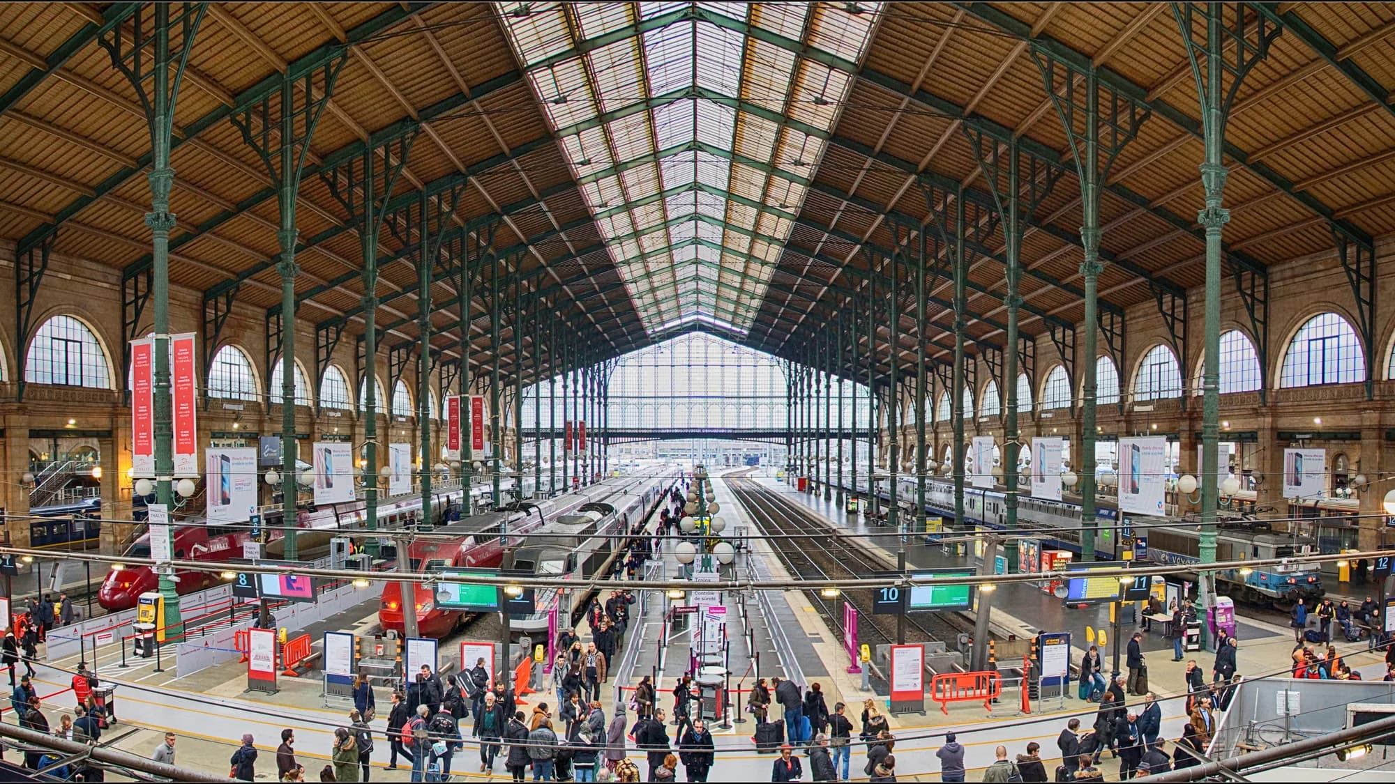 Rail advertising at Gare du Nord in Paris, France: hanging banners line the train shed above the platforms, with a red high-speed train and a crowd in the concourse.
