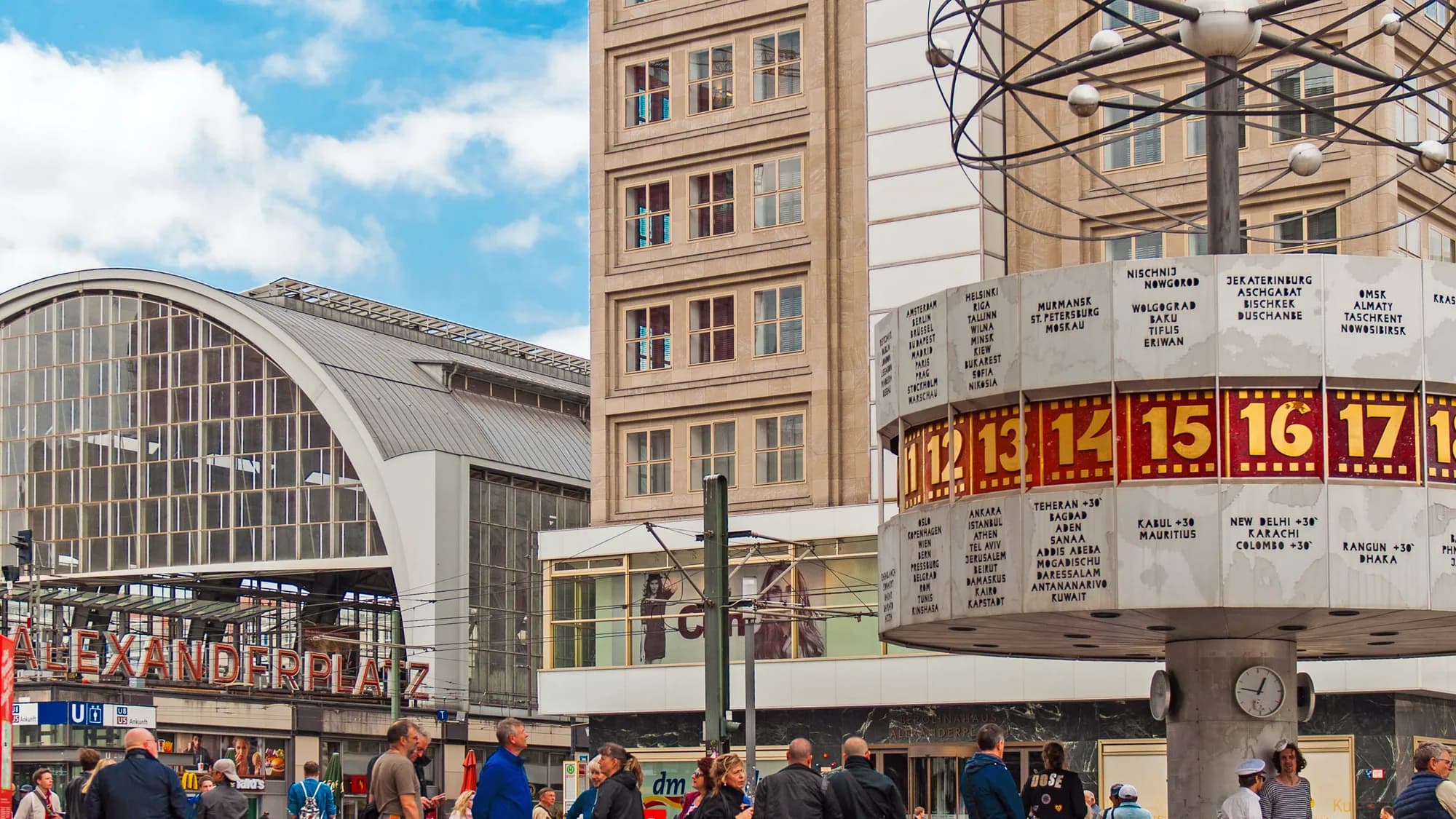 Alexanderplatz in Berlin, Germany: the world time clock stands beside the station's red Alexanderplatz sign, with a crowd in the square.