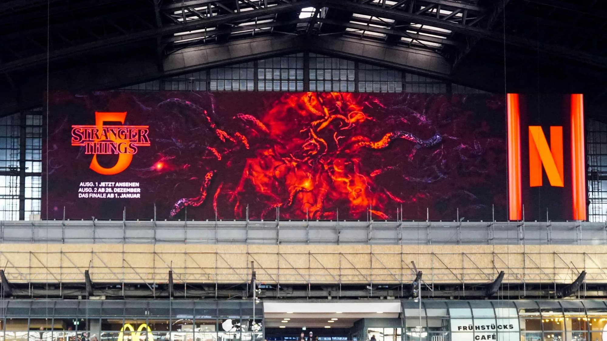 Digital billboard advertising at Hamburg Hauptbahnhof in Germany: a Netflix screen for Stranger Things 5 covers the end wall of the hall above a McDonald's.