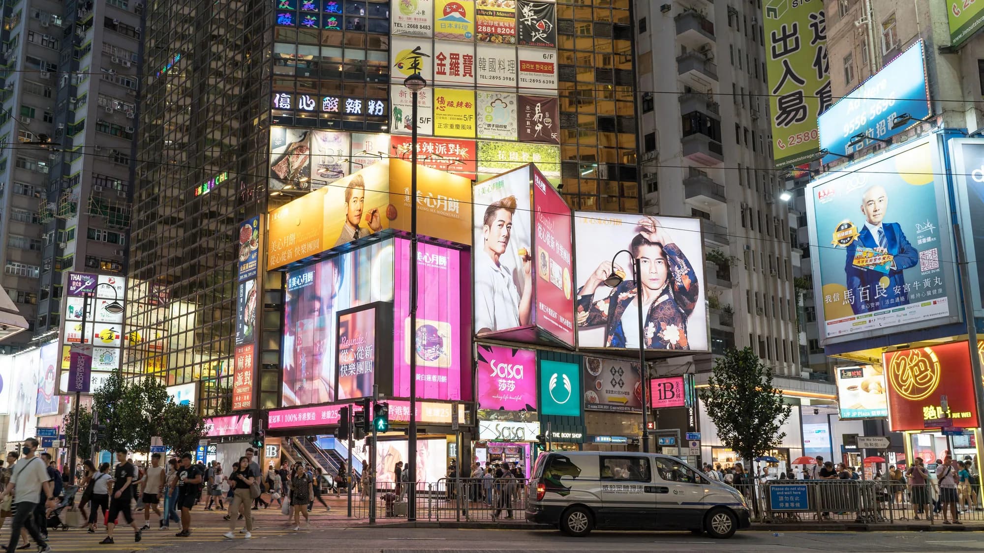 Billboard advertising in Hong Kong: night-time boards cover the towers of a crowded shopping street, including bakery ads and portrait screens.