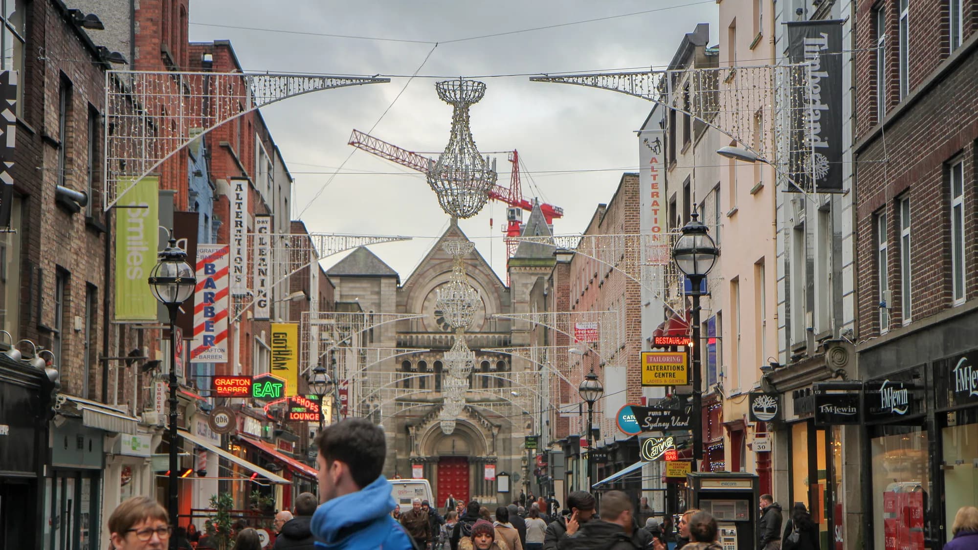 Grafton Street in Dublin, Ireland: Christmas lights span the shopping street above a crowd, with shop signs and a stone church ahead.