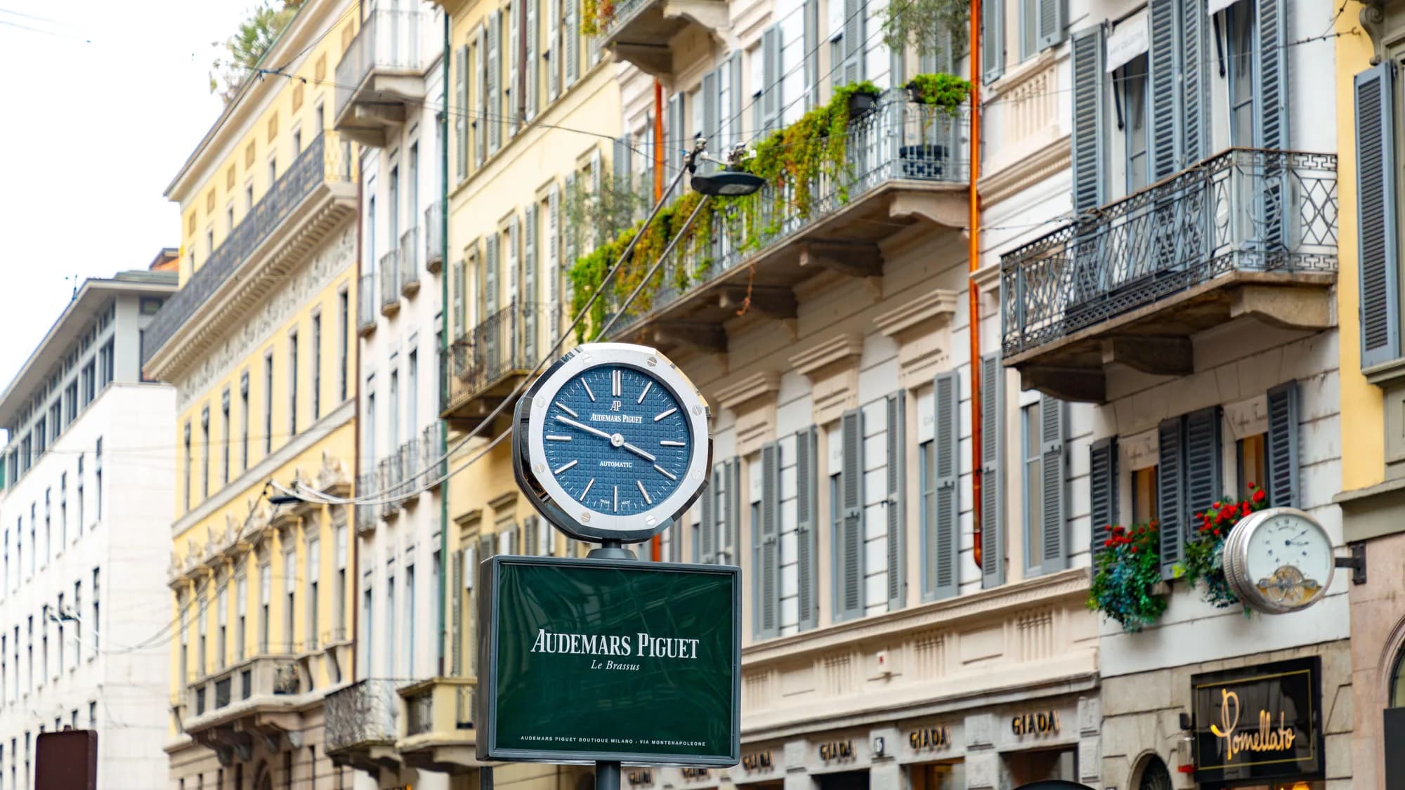 Street furniture in Milan's Quadrilatero della Moda, Italy: an Audemars Piguet street clock stands on the corner, with Pomellato beside it.