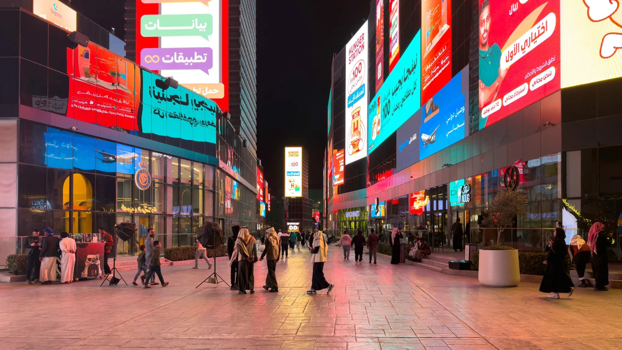 Digital billboard advertising in Saudi Arabia: STC, AliExpress and other screens line both sides of a pedestrian shopping street at night.