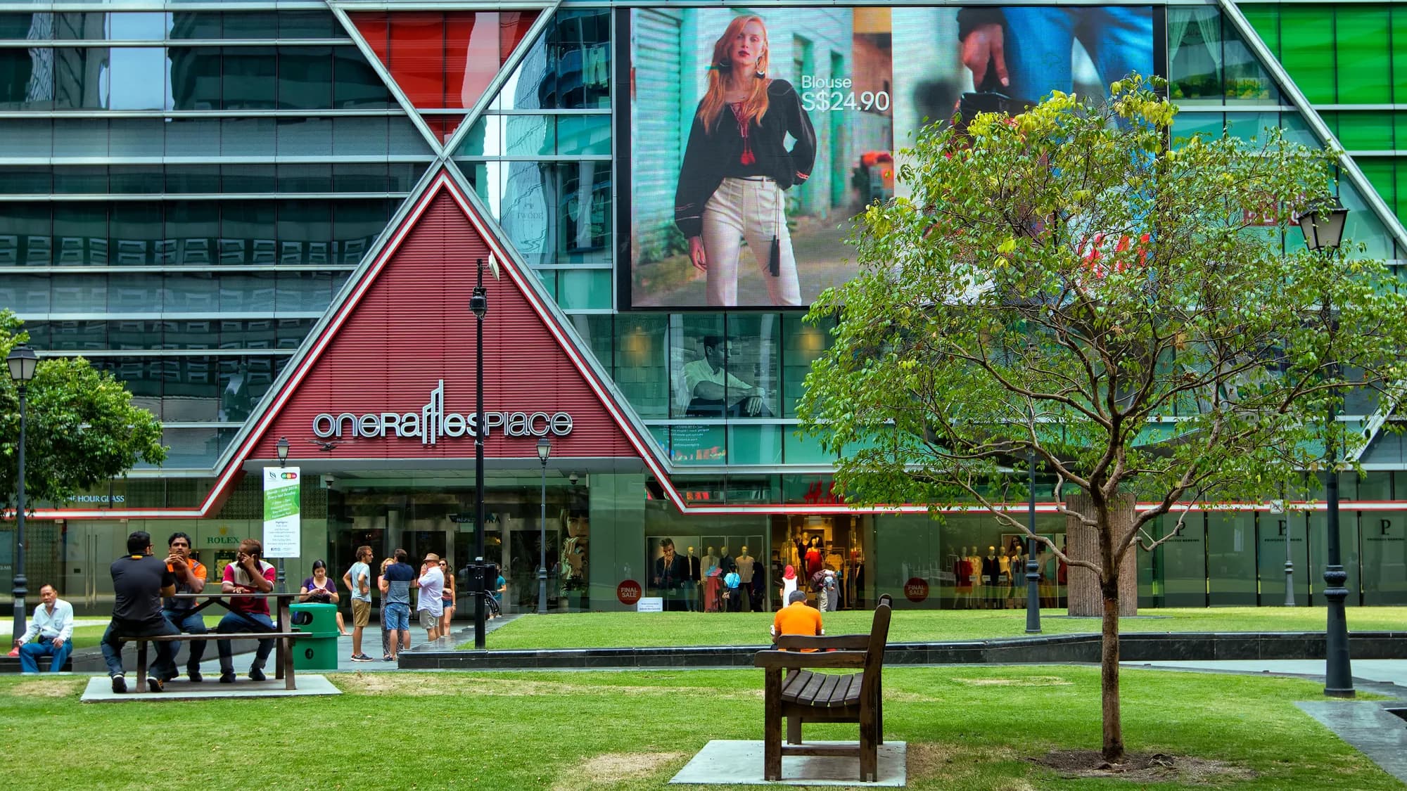 Building banner advertising at Raffles Place in Singapore: a skincare advertisement covers the glass front of One Raffles Place, with another poster on the lawn below.