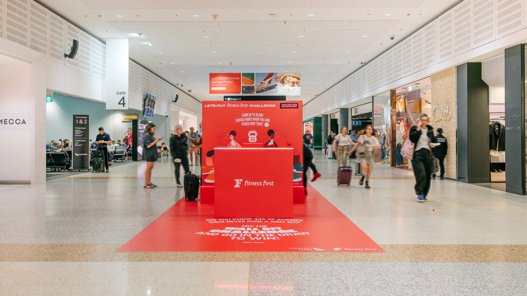 A brand activation at an Australian airport: a red Fitness First stand offers the Let's Put Fitness First challenge, with Qantas points, beside Gate 4.