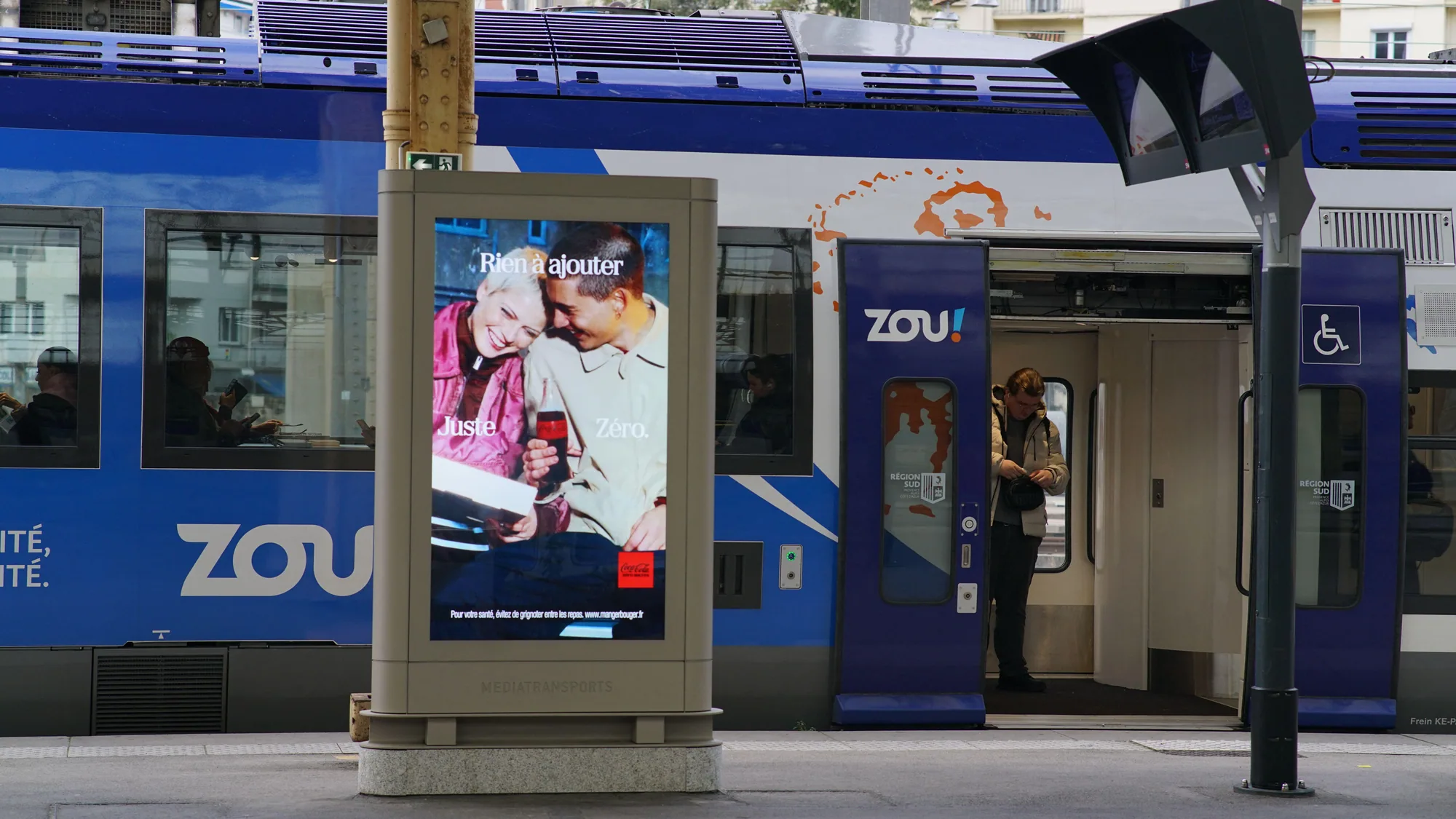 Rail advertising in France: a Coca-Cola Zero poster reading Rien à ajouter, Juste Zéro stands beside a blue ZOU! train.