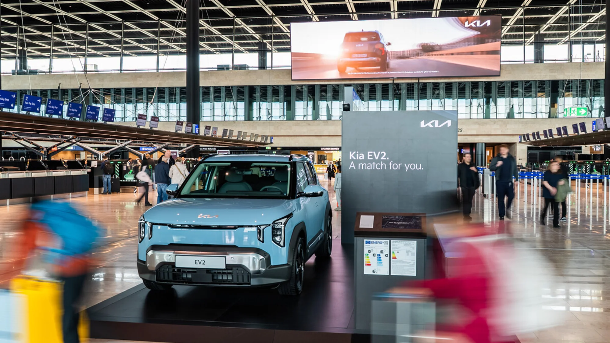 A Kia EV2 brand activation in a German airport terminal shows the car on a plinth, with the line A match for you, and a Kia screen above it.