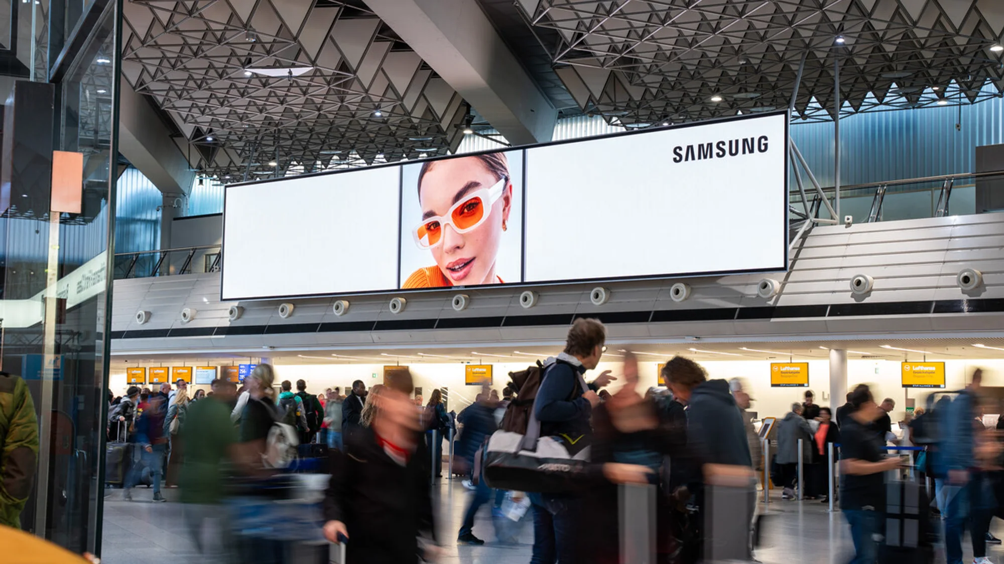 Airport advertising in Germany: a wide Samsung screen in a departure hall shows a woman in orange sunglasses, above yellow flight boards.