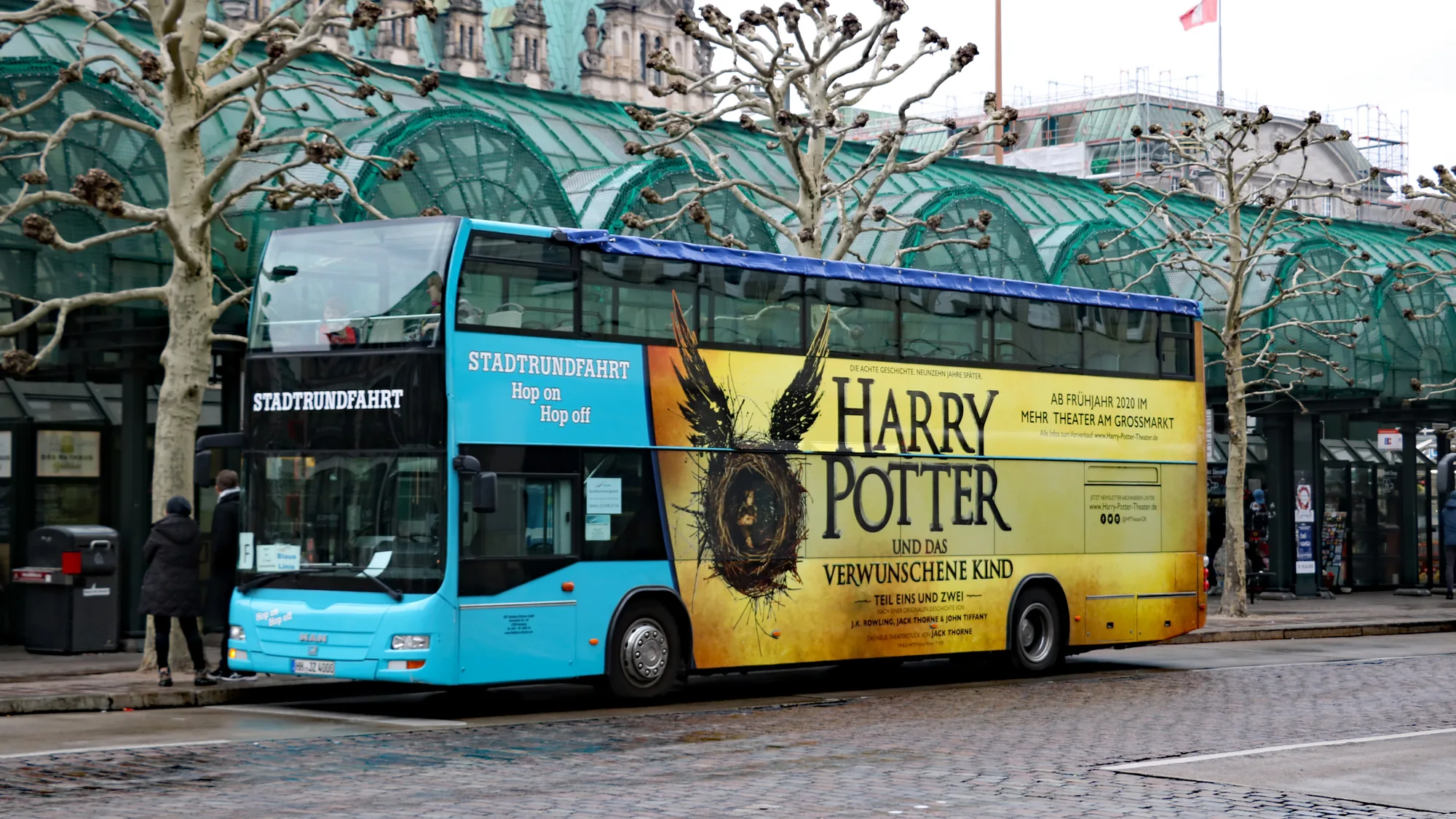Bus advertising in Hamburg, Germany: a turquoise Stadtrundfahrt double-decker carries a yellow Harry Potter und das verwunschene Kind wrap beside a glass-roofed station.