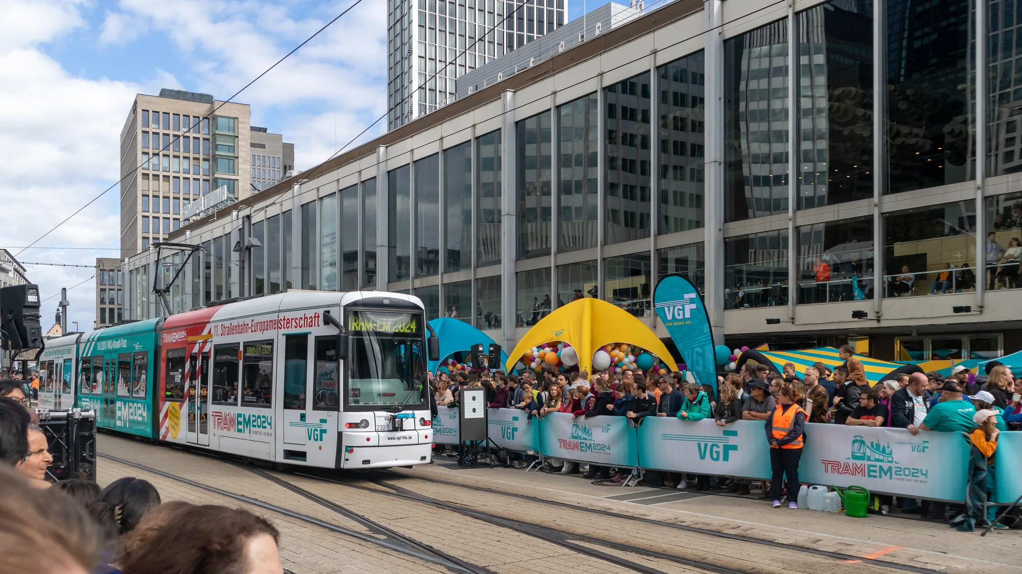 Tram advertising in Frankfurt, Germany: a white and teal tram for the 11. Straßenbahn-Europameisterschaft reads TRAM EM 2024, beside VGF tents and a crowd.