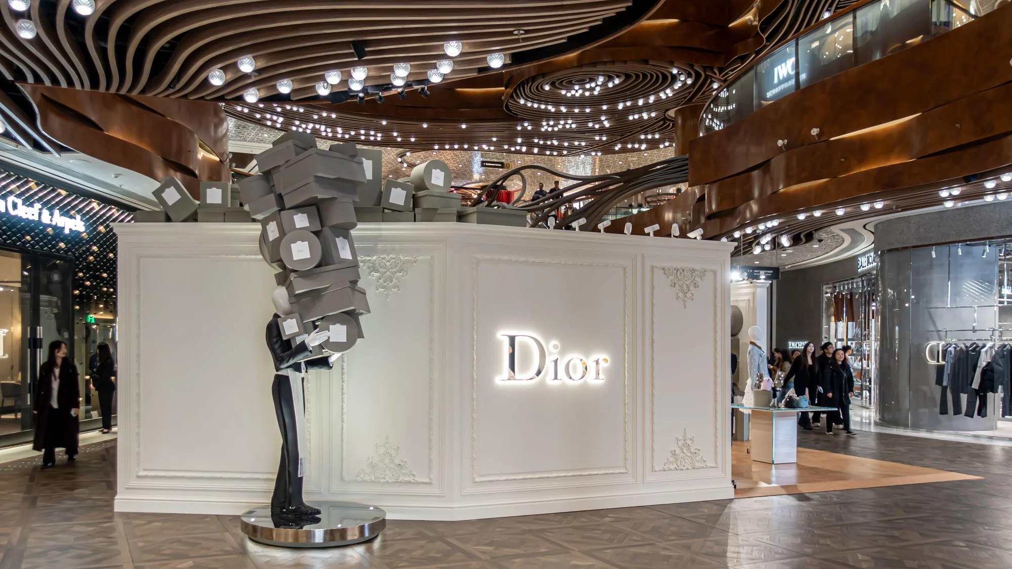 A Dior brand activation in a Hong Kong mall: a mannequin carries a tall stack of grey hat boxes beside an illuminated Dior wall.
