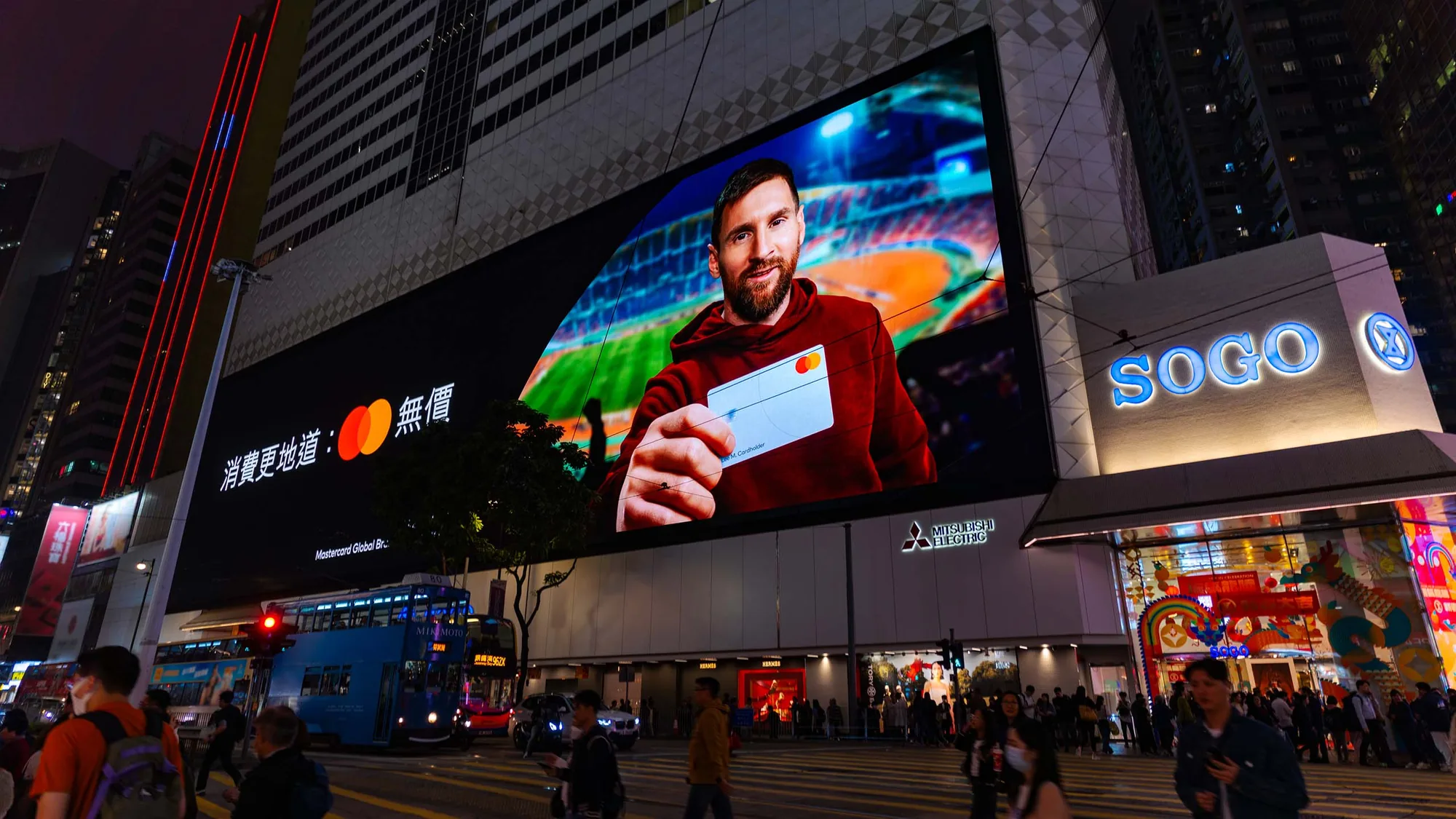 Digital out-of-home advertising in Hong Kong: a night billboard shows Lionel Messi holding a Mastercard, beside the lit SOGO store and a tram.
