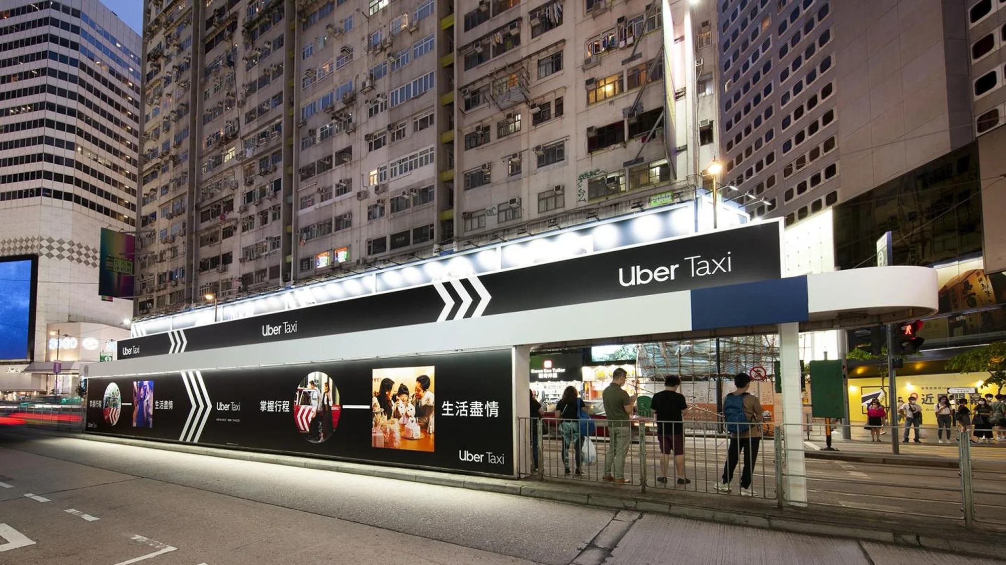 Street furniture advertising in Hong Kong: an Uber Taxi wrap covers a lit tram shelter at night, with people waiting beneath the towers.