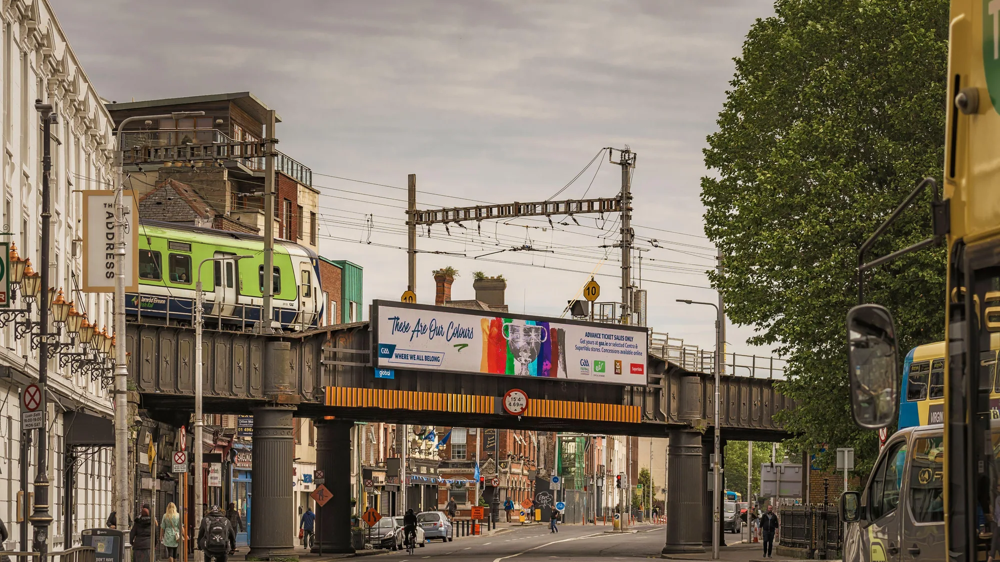 Bridge banner advertising in Ireland: a railway bridge carries a GAA banner reading These are our colours, while a green train crosses above the street.