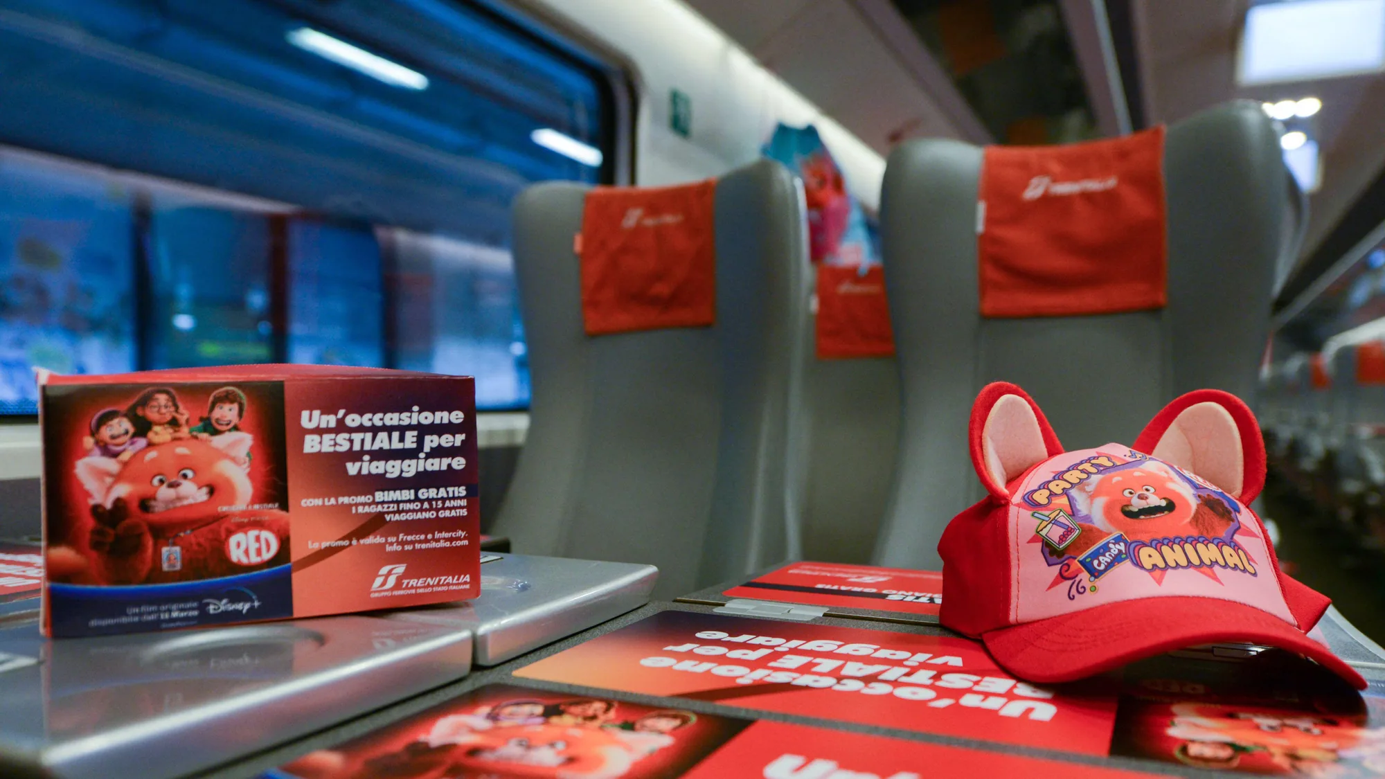 Rail advertising in Italy: a Trenitalia table holds Turning Red cards reading Un'occasione bestiale per viaggiare, plus a red panda hat.