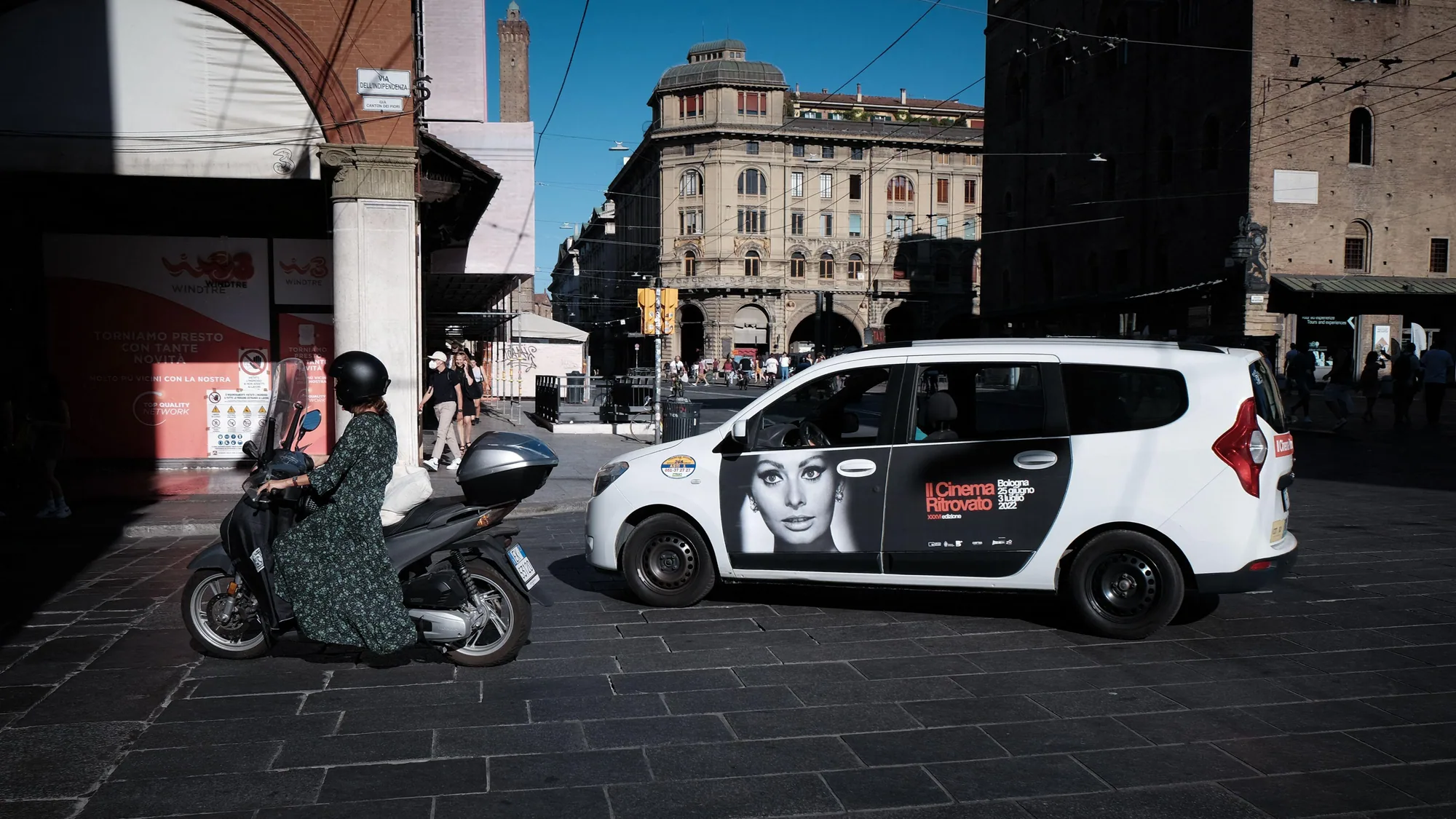 Taxi advertising in Bologna, Italy: a white taxi carries a Cinema Ritrovato door advert of Sophia Loren while a rider in a dress passes beside it.