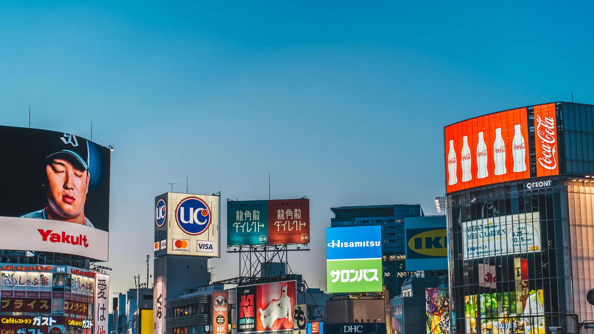 Billboard advertising in Japan: screens over a city crossing include Yakult, Coca-Cola, Hisamitsu Salonpas and a UC card, beside the QFRONT building.