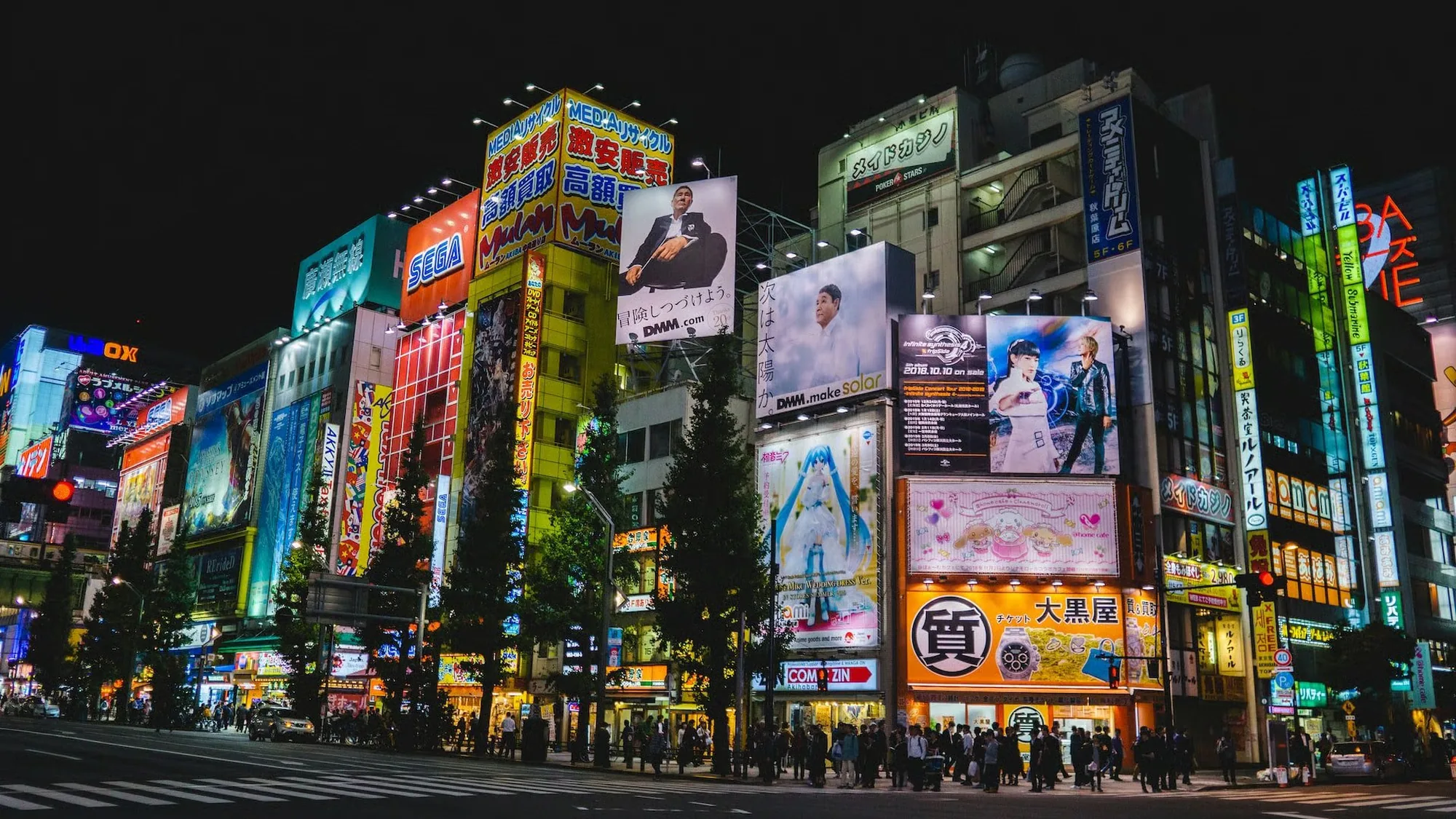 Building banner advertising in Japan: neon signs for SEGA and DMM.com, plus a large Hatsune Miku advertisement, rise over a crowded street at night.