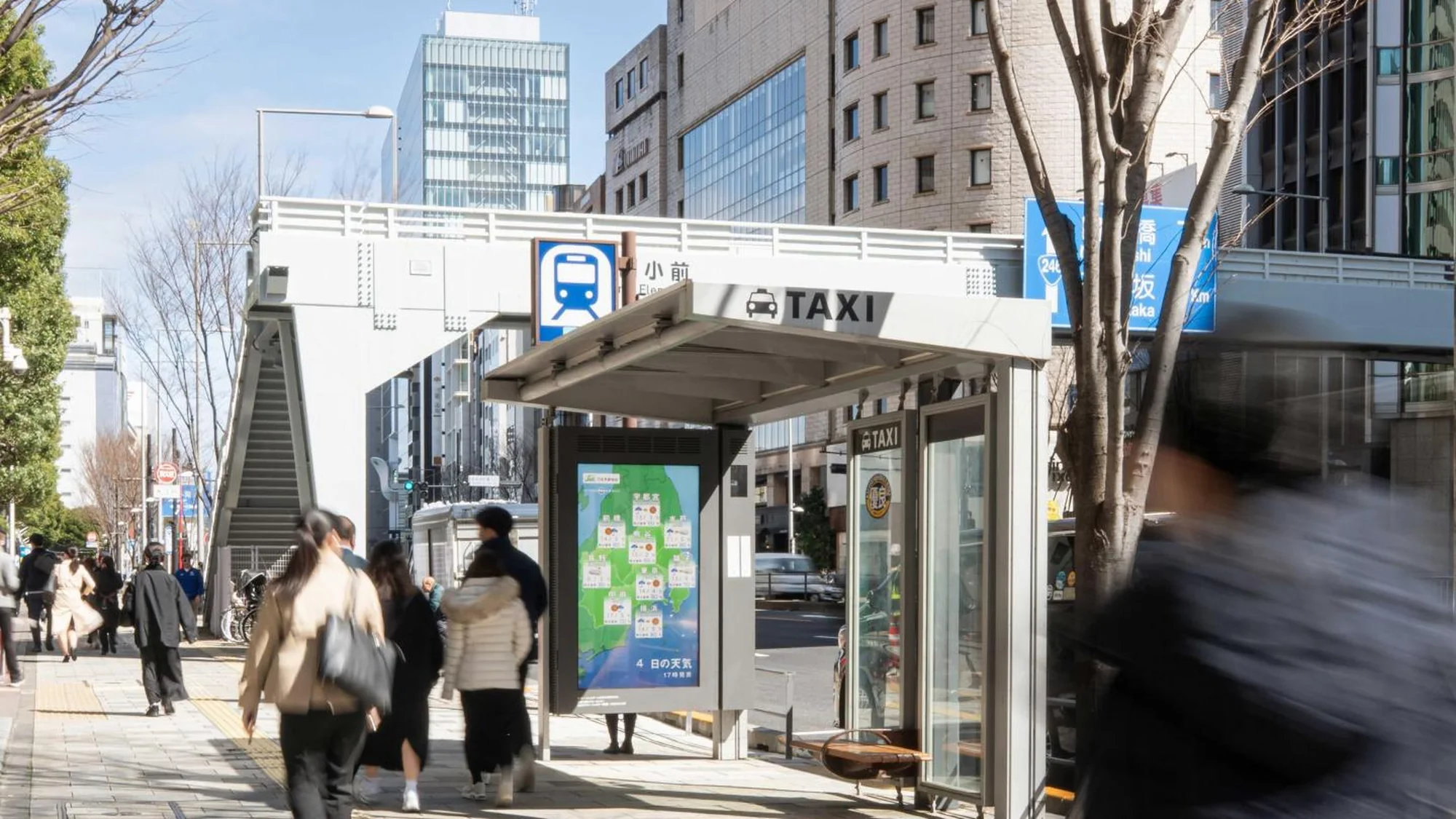 Programmatic digital advertising in Japan: a screen on a taxi shelter shows a weather map, beneath a station sign and platform stairs.