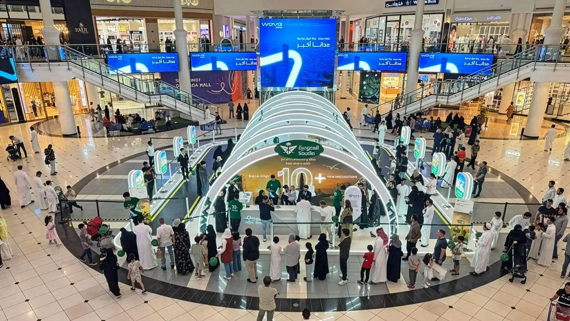 A Saudia brand activation in a Saudi shopping mall: a stand marked 10+ sits under a lit arch and blue digital screens.