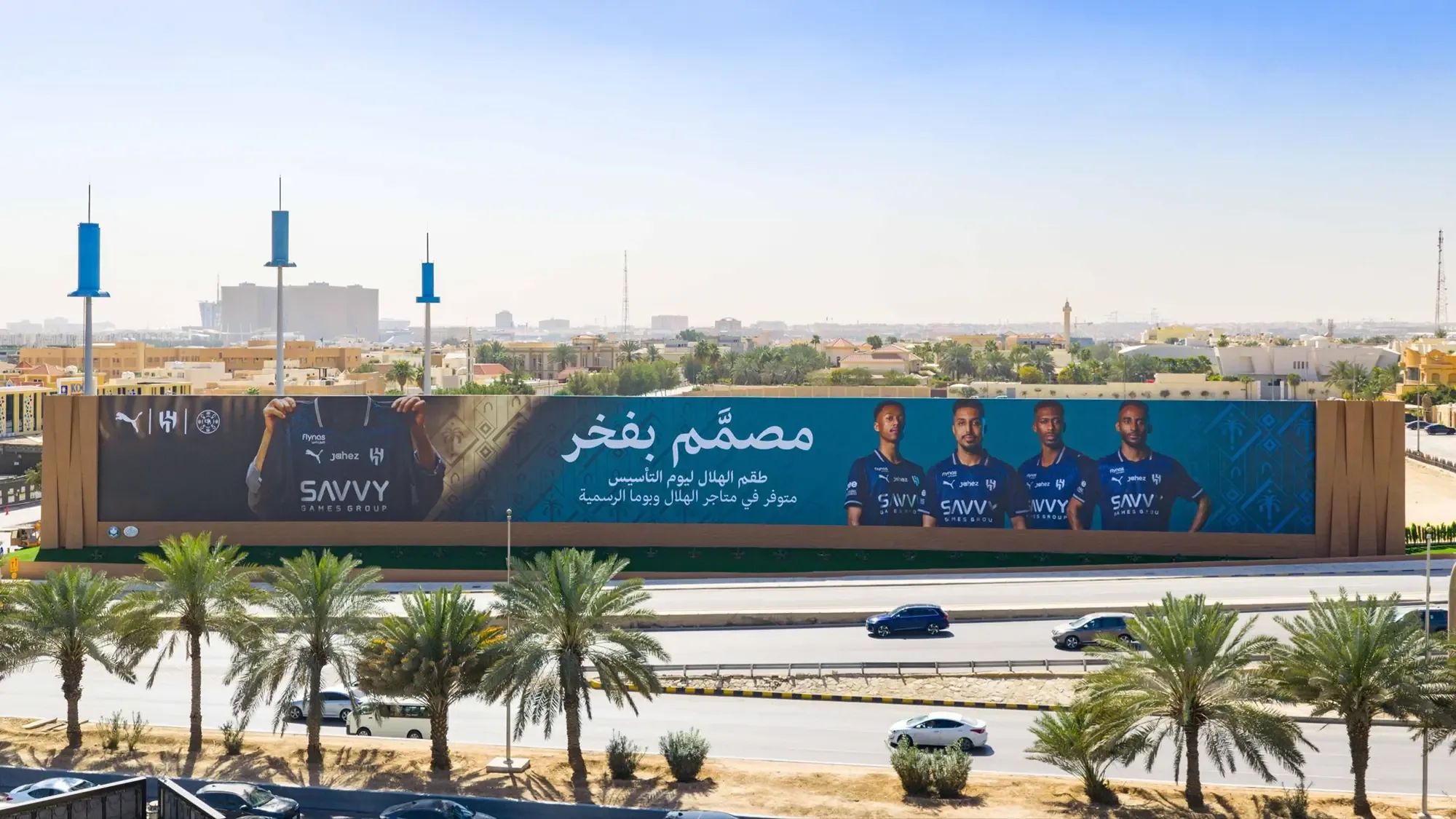 Large format banner advertising in Saudi Arabia: a long roadside banner shows Al Hilal players in Savvy Games Group shirts under an Arabic headline.