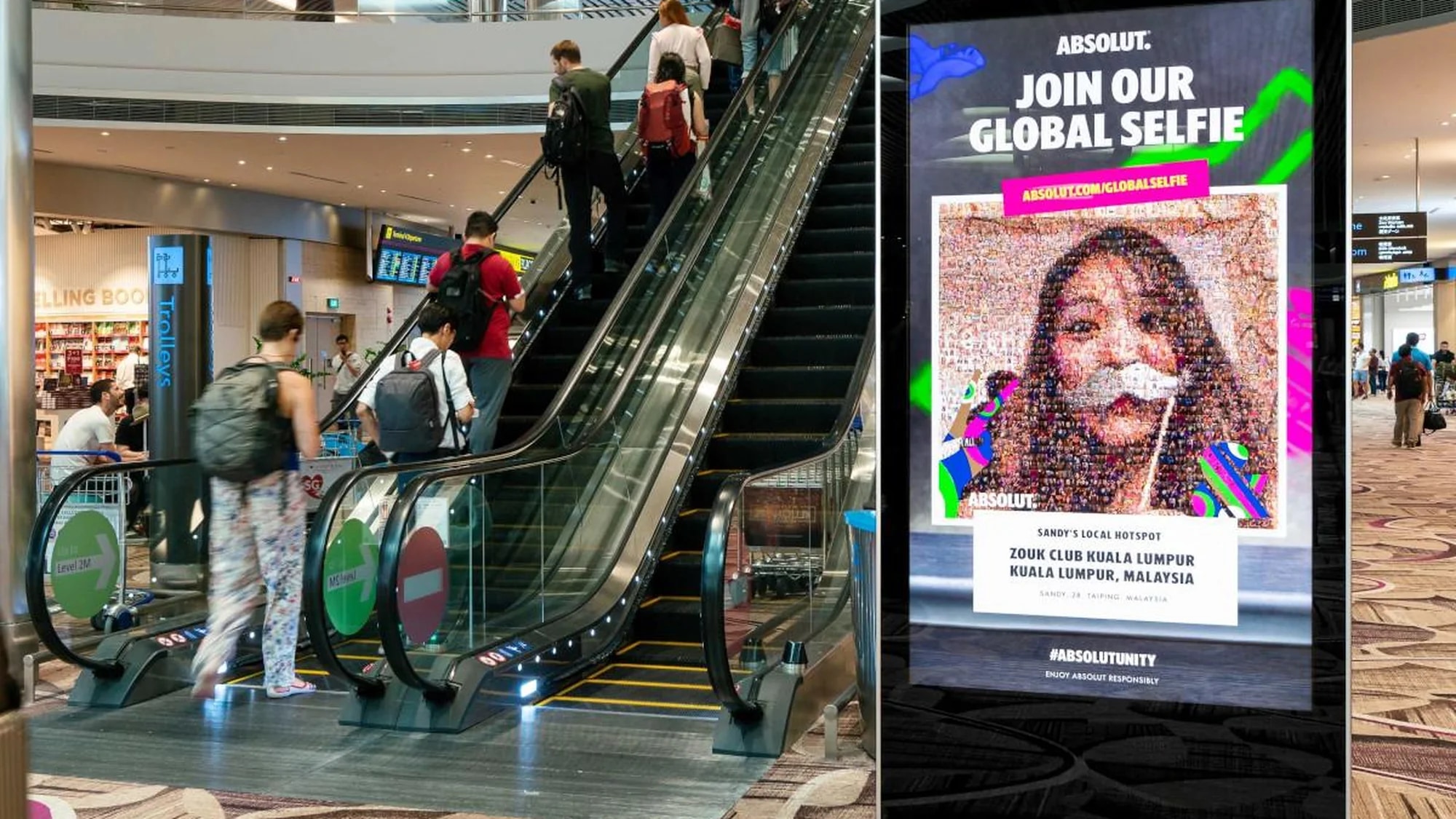 Digital out-of-home advertising beside airport escalators in Singapore: an Absolut screen shows Join our global selfie, with a mosaic portrait for Zouk Club in Kuala Lumpur.