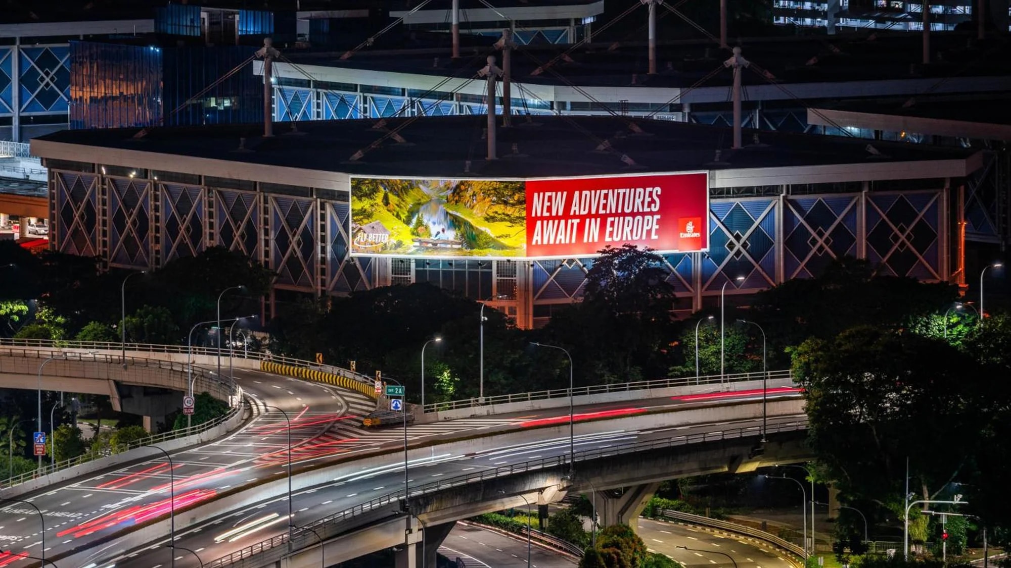 Billboard advertising in Singapore: a large night board reads New adventures await in Europe, on a curved stadium building above a highway.