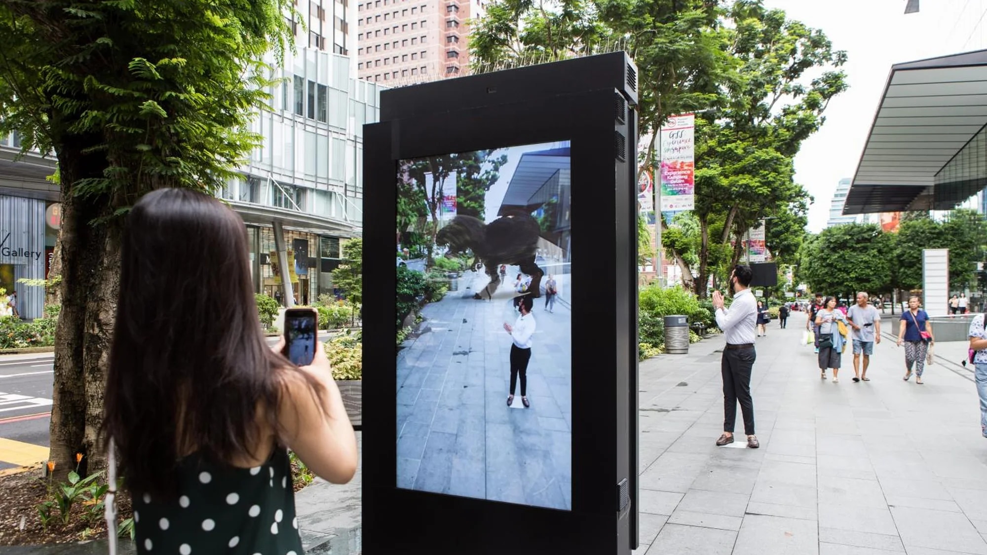 Street furniture advertising in Singapore: people on a pavement film a street screen that shows a dinosaur standing over a woman.