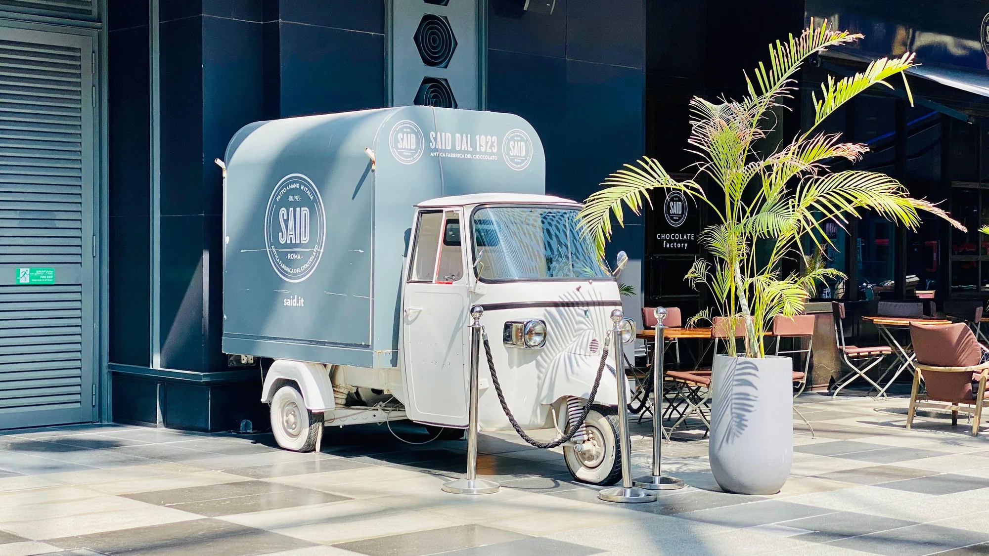 A brand activation: a three-wheeled Said dal 1923 van is parked outside a chocolate shop, with cafe tables beside it.