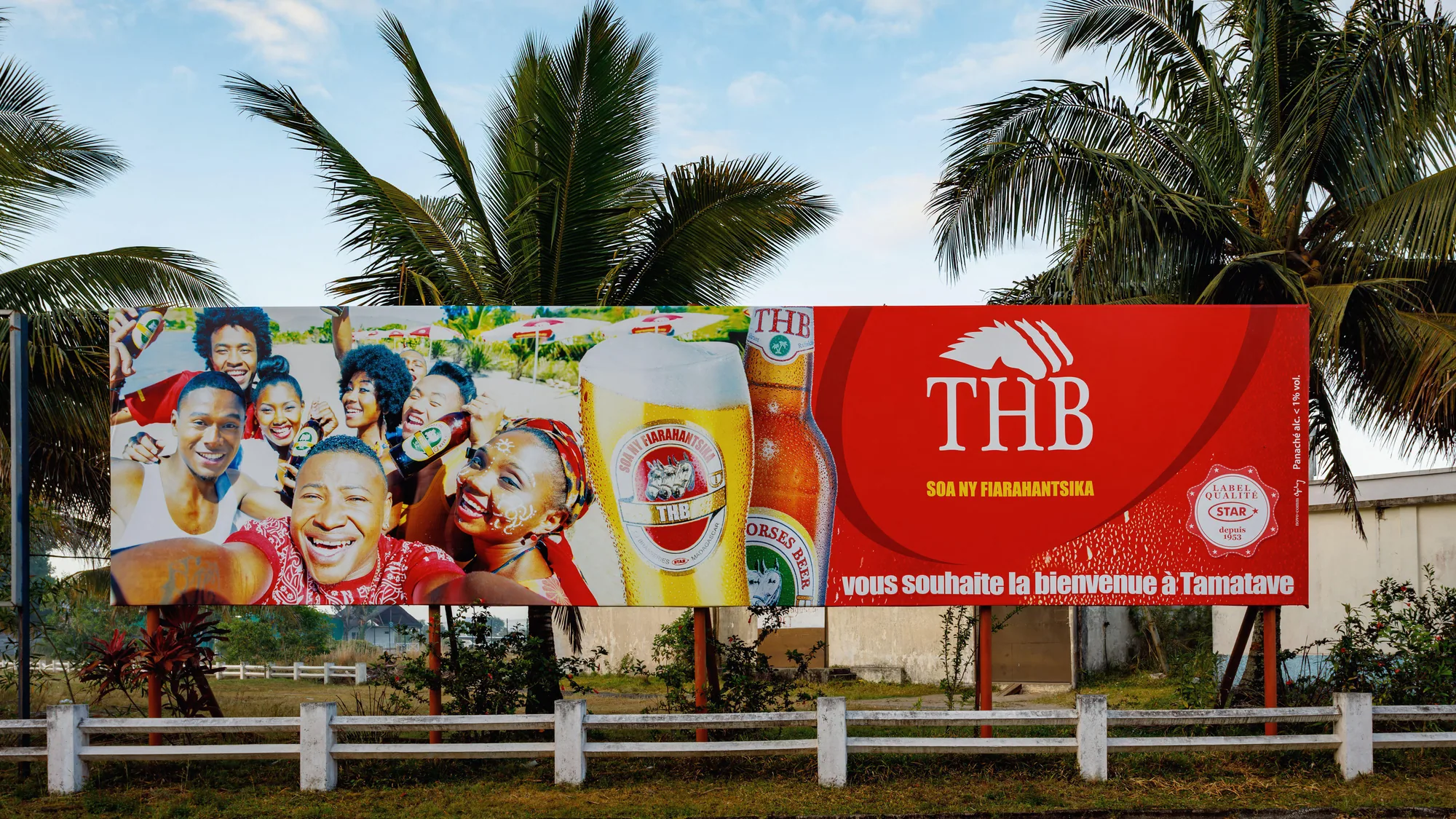 Large format billboard advertising in Tamatave, Madagascar: a board for THB Three Horses Beer welcomes visitors, with palm trees behind it.