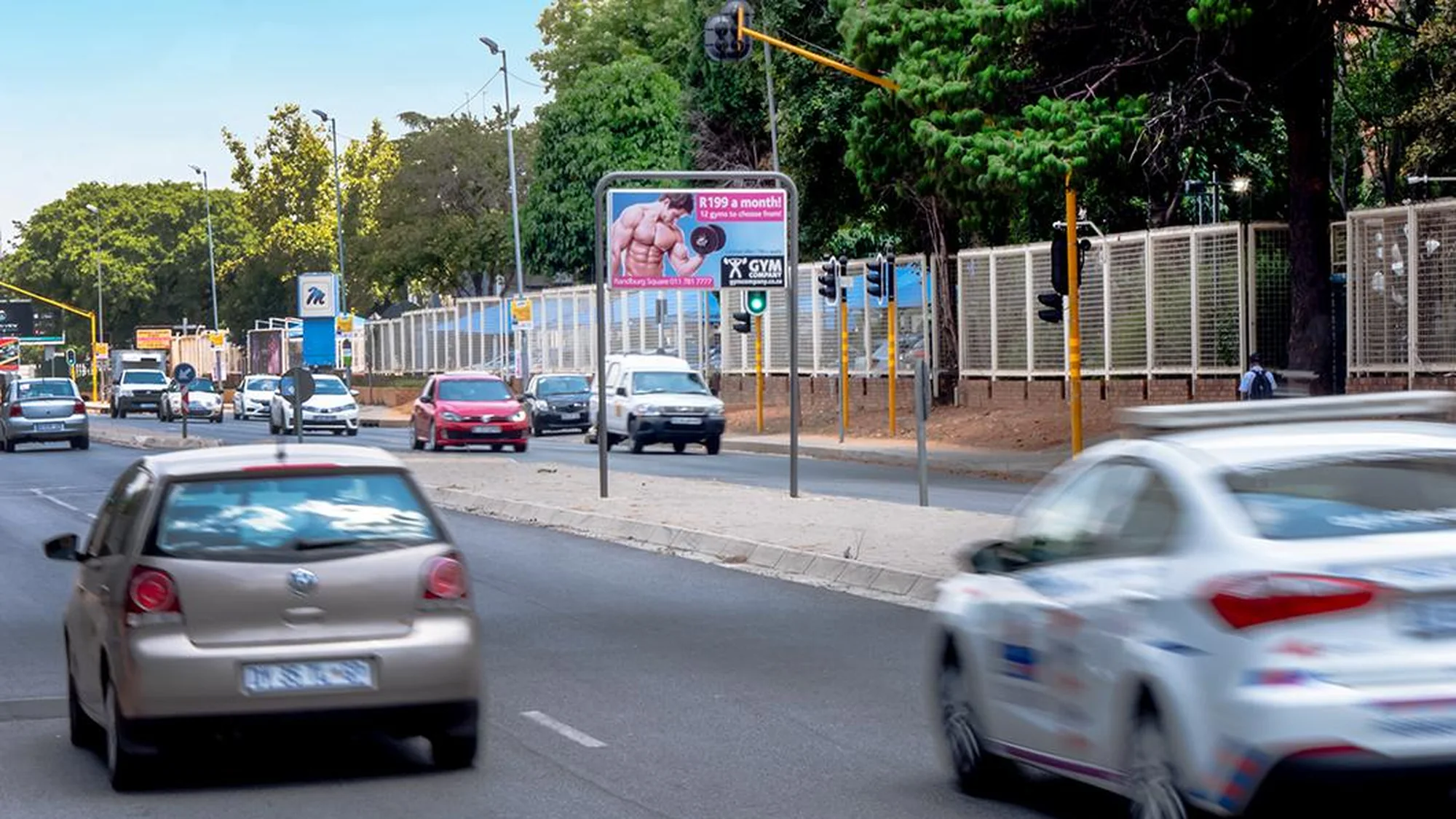Street furniture advertising in South Africa: a roadside poster advertises X Gym at R199 a month, above traffic at a junction.