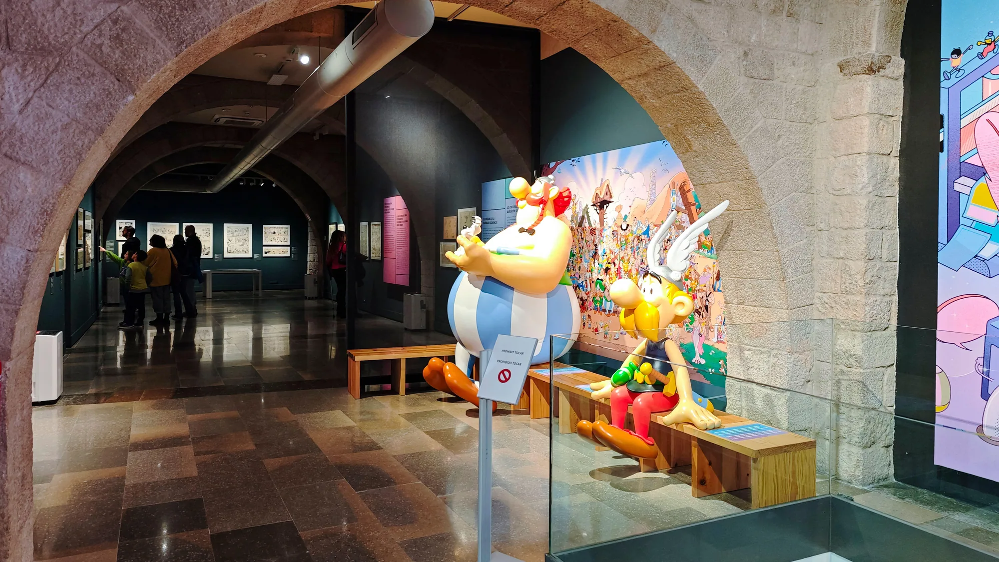 A brand activation in Spain: Asterix and Obelix statues stand in a stone-arched exhibition hall, with visitors and a comic mural behind them.