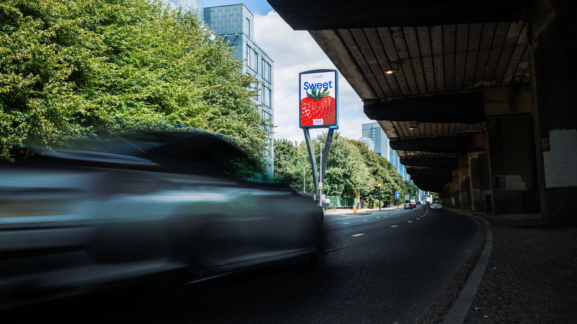 Billboard advertising in the United Kingdom: a Tesco board showing a strawberry and the word Sweet stands beside a road under a flyover.