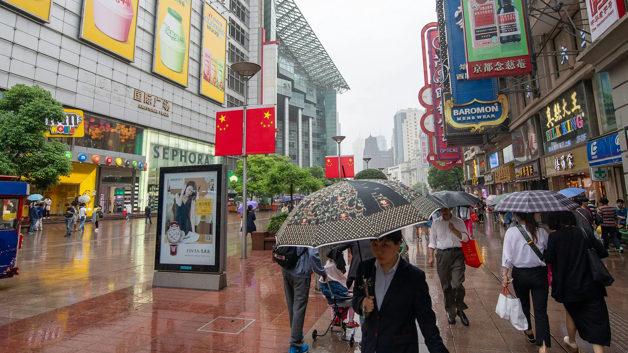 Nanjing Road in Shanghai, China: umbrella-carrying shoppers pass Sephora, Chinese flags and hanging shop signs in the rain.