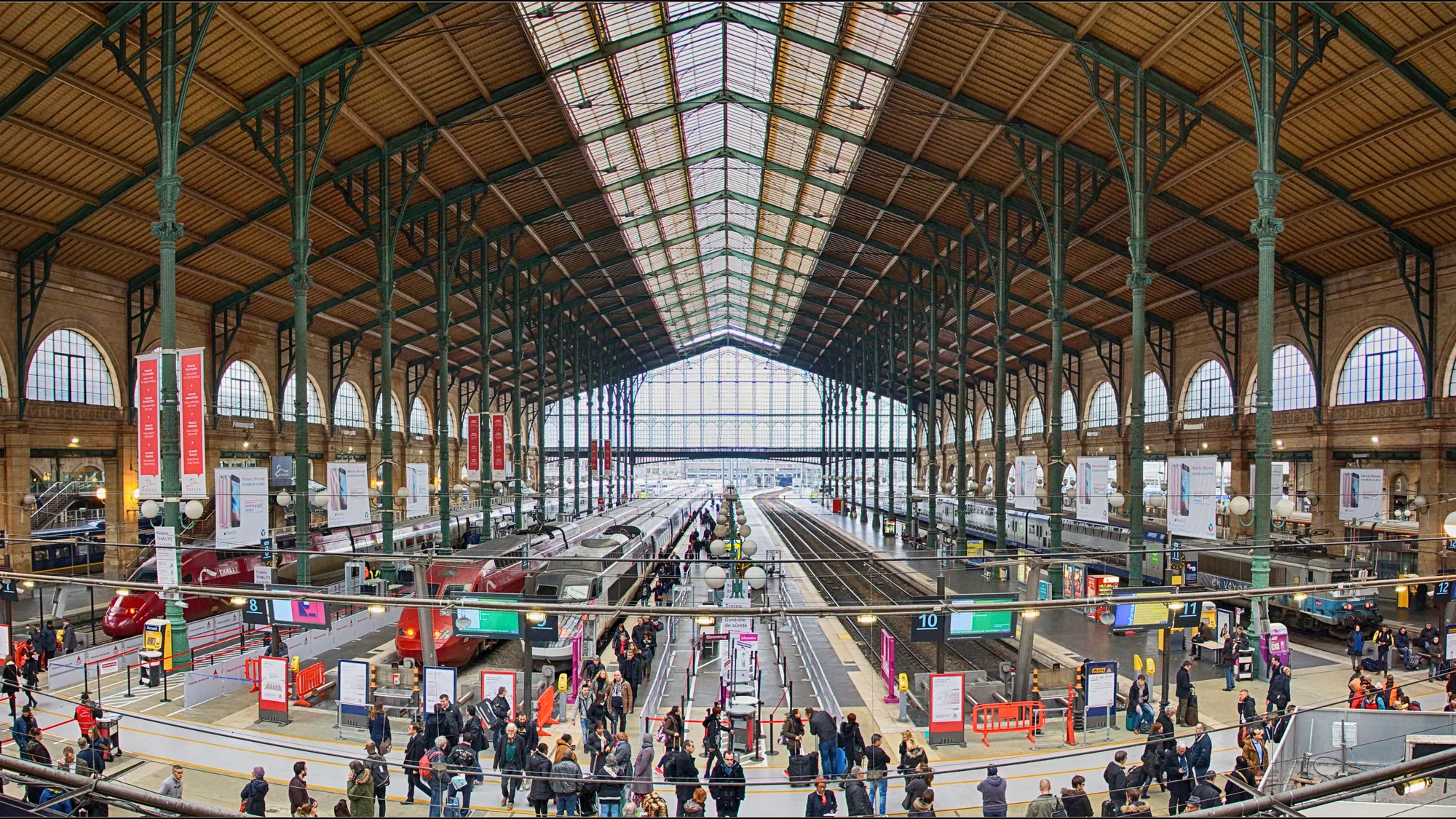 Rail advertising at Gare du Nord in Paris, France: hanging banners line the train shed above the platforms, with a red high-speed train and a crowd in the concourse.