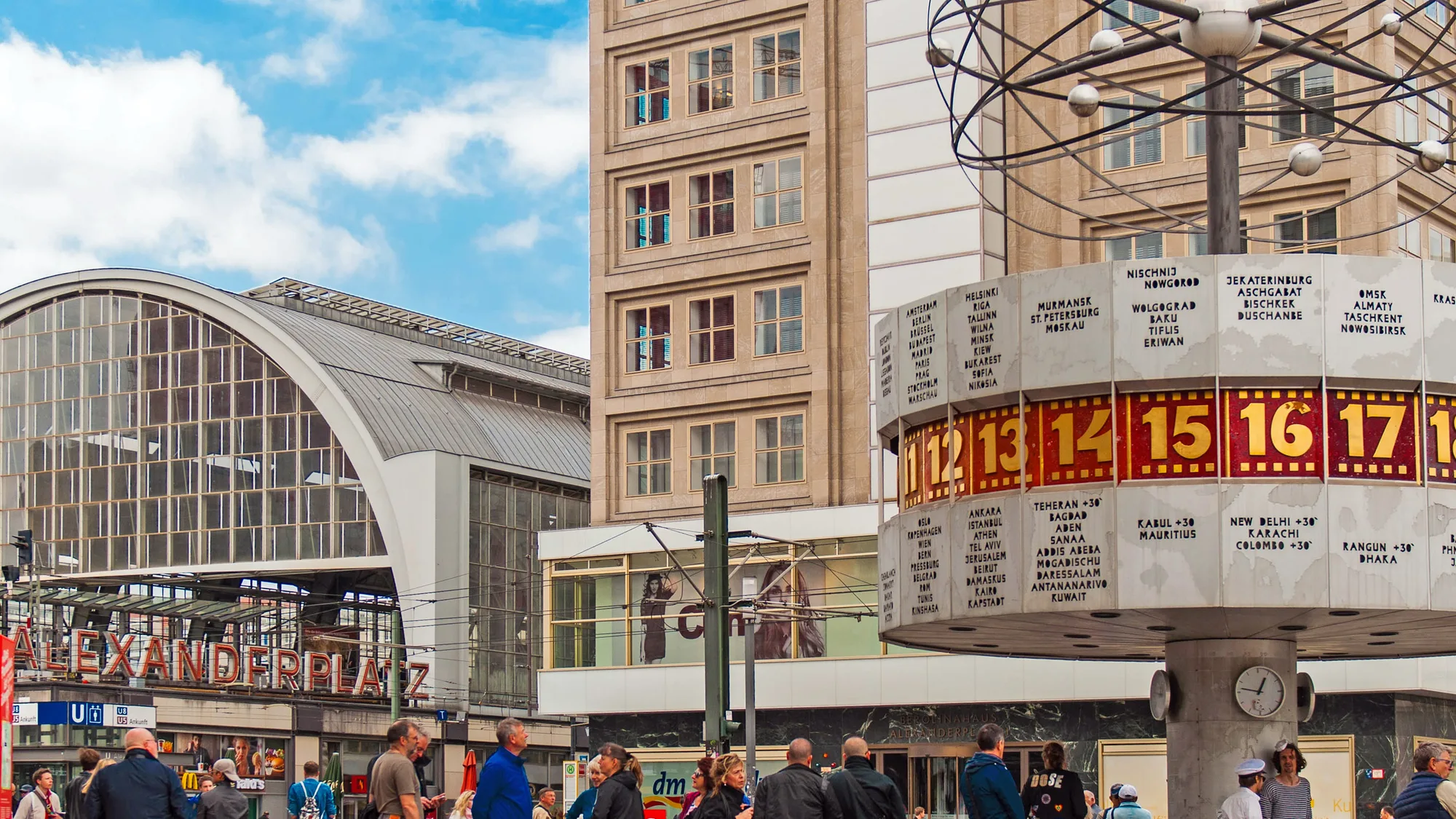 Alexanderplatz in Berlin, Germany: the world time clock stands beside the station's red Alexanderplatz sign, with a crowd in the square.