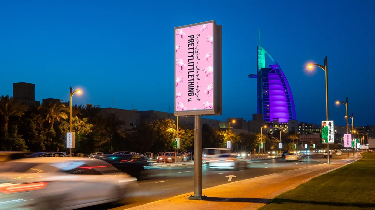 Street furniture advertising in Dubai, in the UAE: a PrettyLittleThing roadside screen glows at dusk, with the Burj Al Arab lit purple behind it.