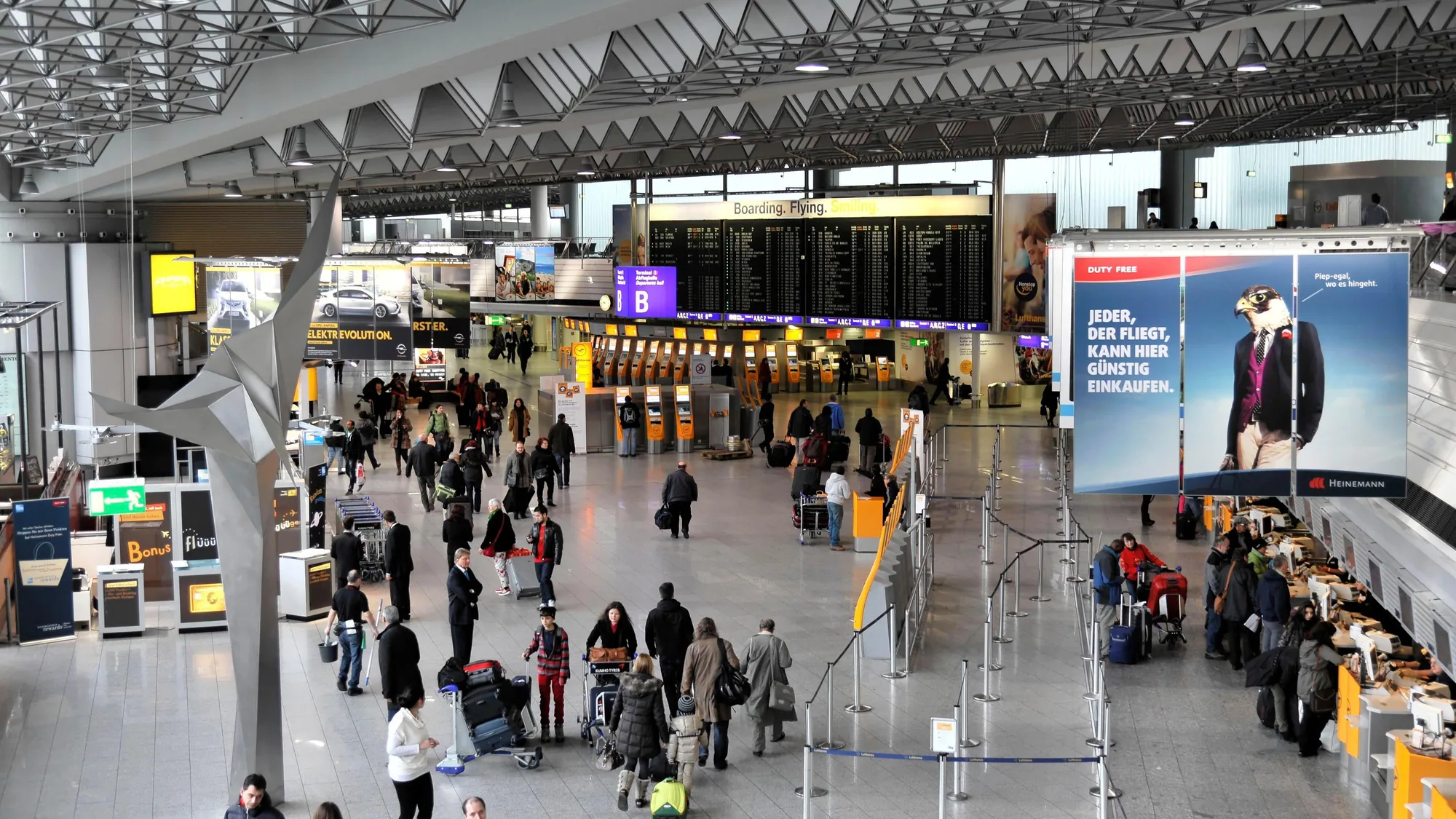 Airport advertising at Frankfurt Airport in Germany: travellers queue in the departure hall beneath the flight boards and a Heinemann duty-free banner.