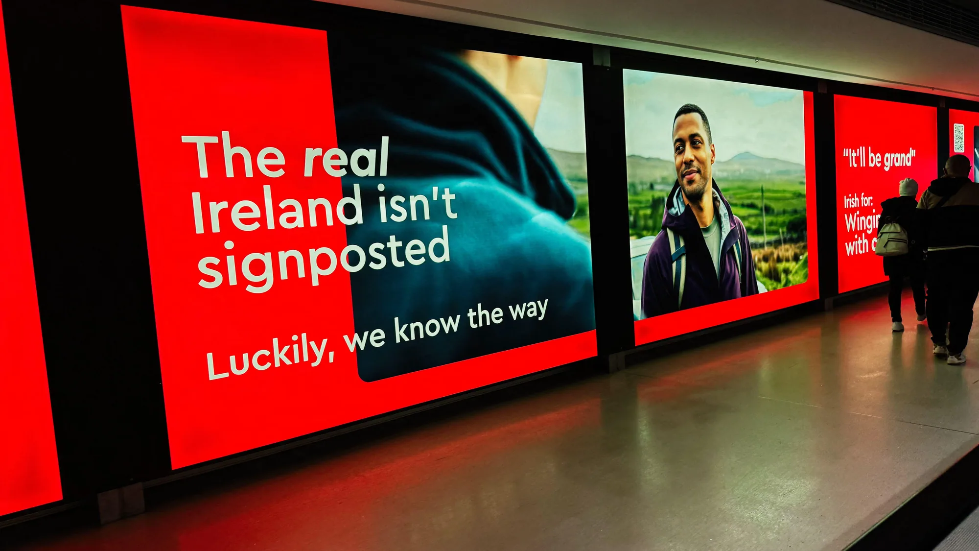 Airport advertising at Dublin Airport in Ireland: a red poster reading it'll be grand lines a moving walkway, beside FREENOW taxi screens.
