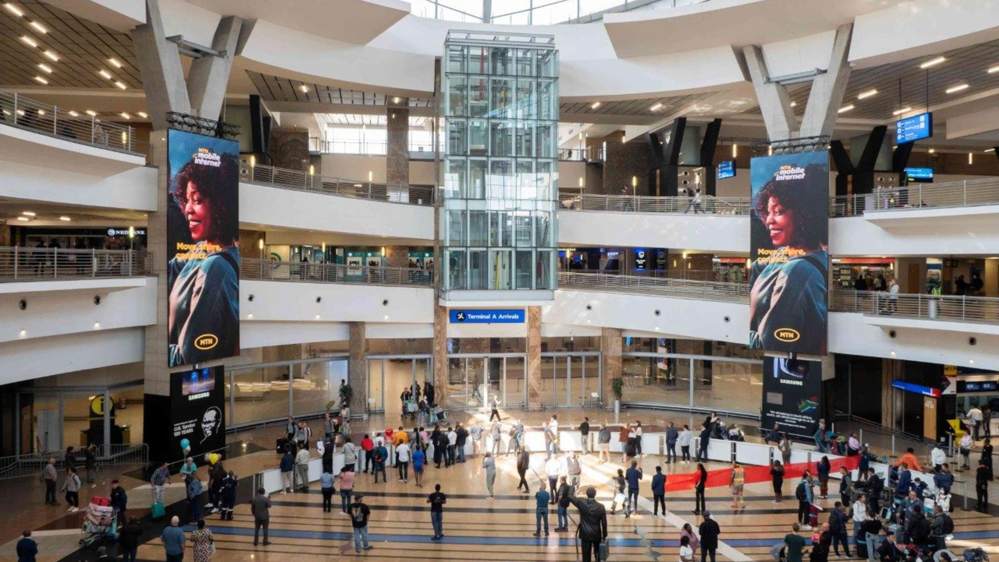 Airport advertising at OR Tambo Airport in Johannesburg: MTN portrait screens hang in the arrivals atrium, with a Terminal A sign below.