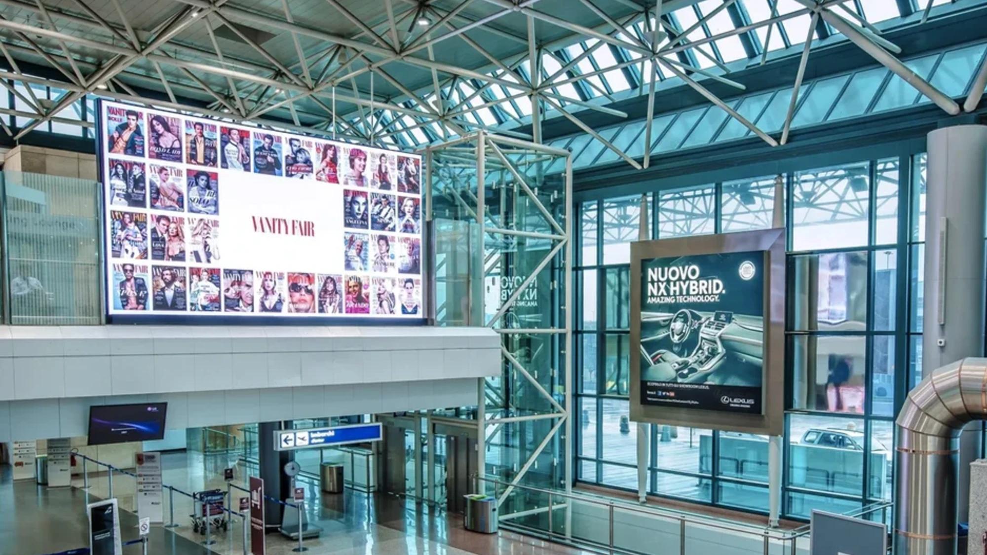Airport advertising in a glass-roofed terminal: Vanity Fair covers and an Italian Lexus hybrid poster face the check-in area.