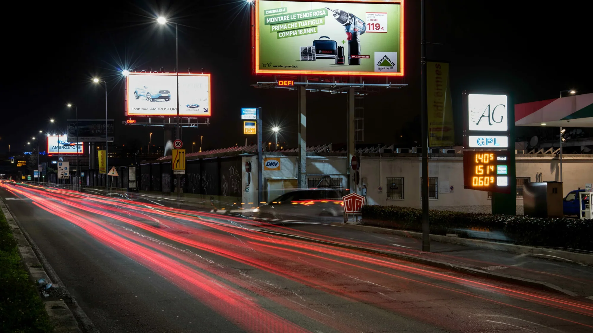 Billboard advertising in Italy: a Leroy Merlin board beside a night-time road shows a drill and the line Montare le tende rosa.