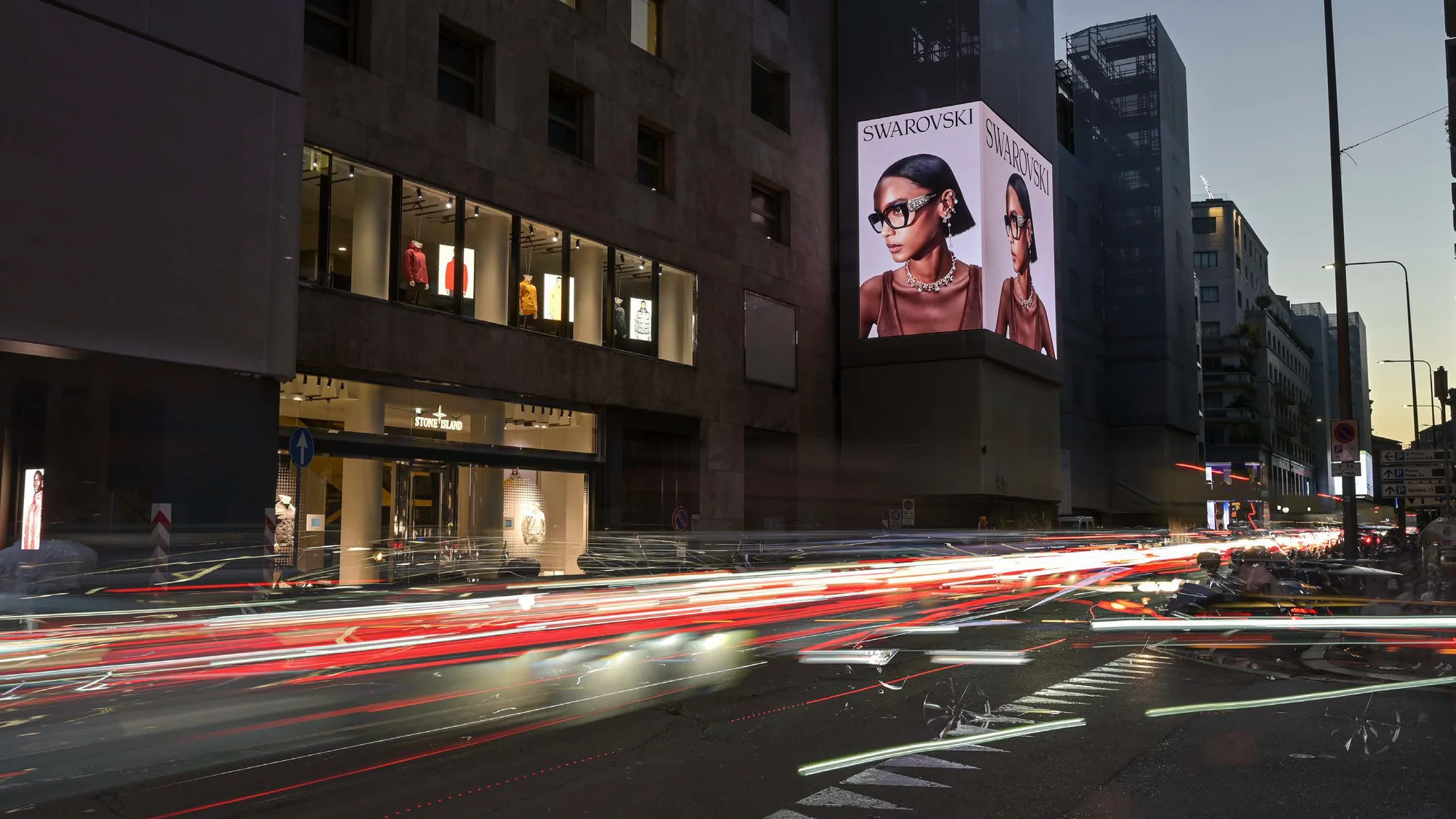 Programmatic digital billboard advertising in Italy: a Swarovski screen of a model in crystal glasses glows above a Stone Island store.