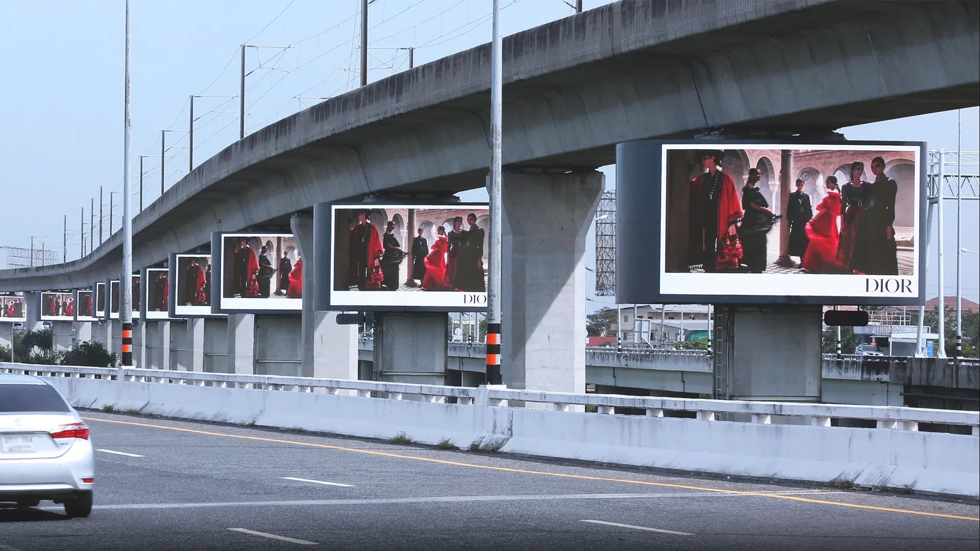 Bridge banner advertising: repeated Dior panels showing models in red and black run along a concrete rail viaduct above a road.
