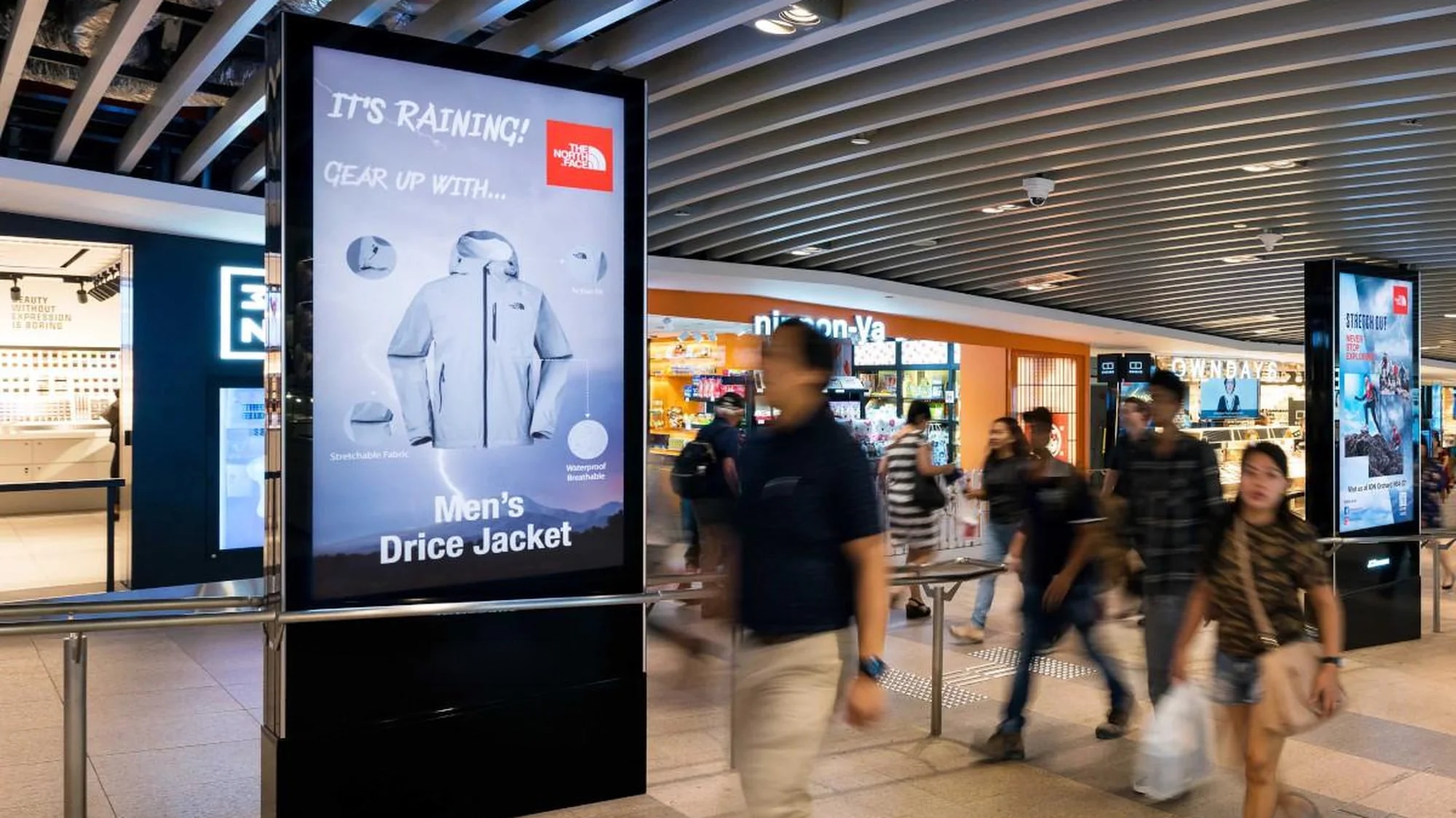 Programmatic digital advertising in a Singapore mall: a North Face screen is headlined It's raining! Gear up, and shows a men's jacket.
