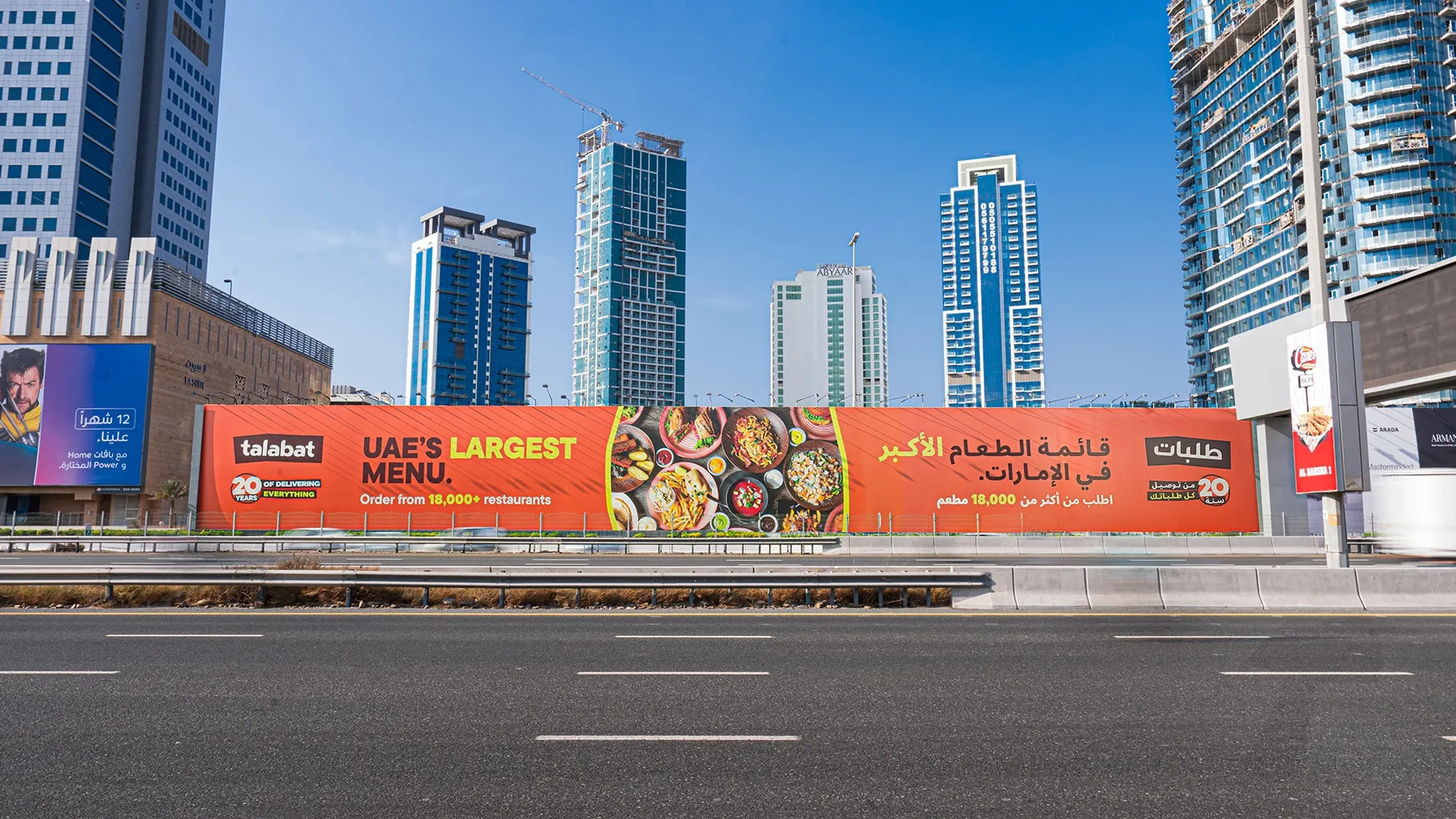 Large format banner advertising in the UAE: an orange talabat hoarding reads UAE's largest menu, order from 18,000+ restaurants, beside a highway and tower blocks.