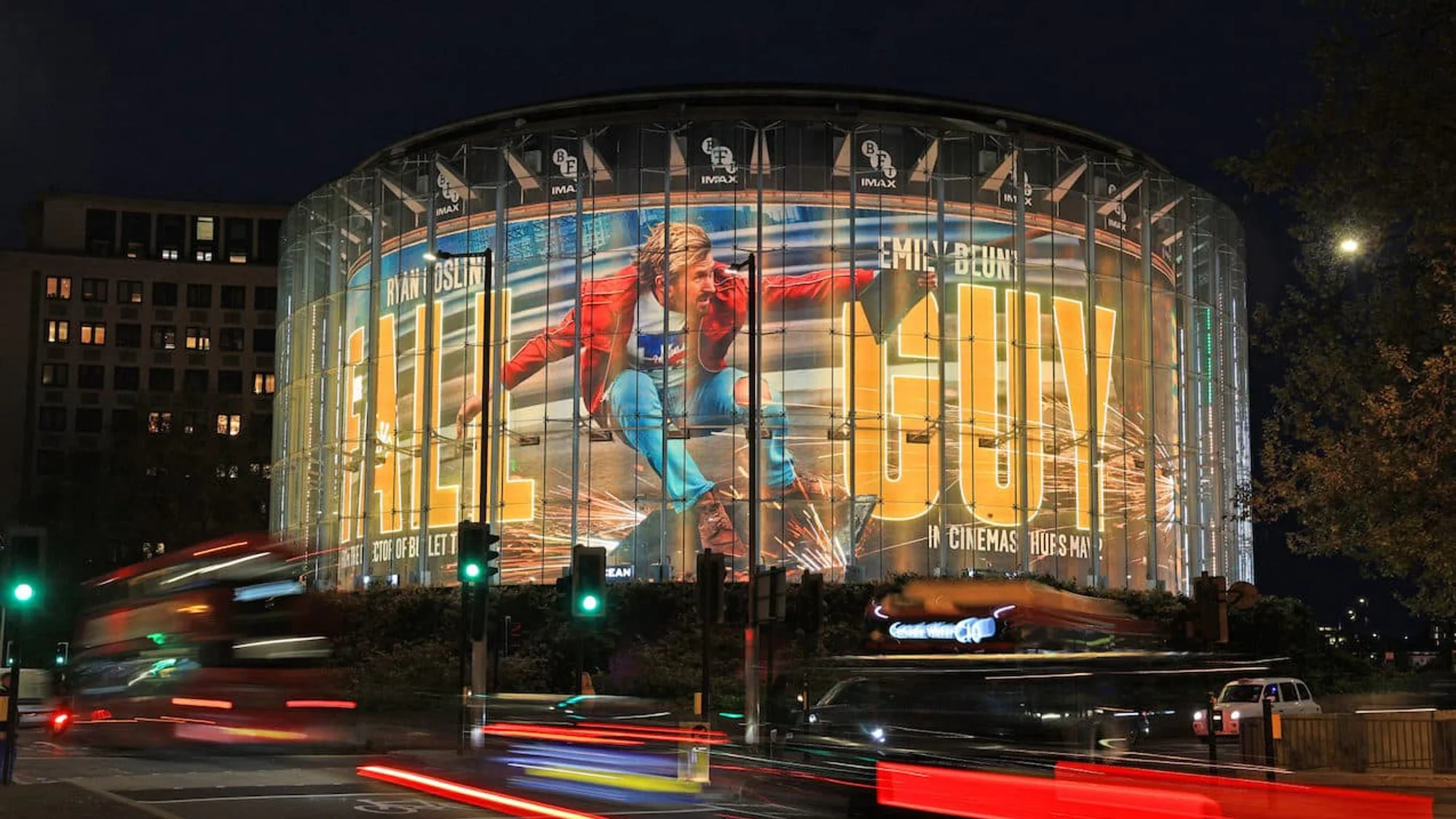 Building banner advertising in London, in the United Kingdom: The Fall Guy, with Ryan Gosling and Emily Blunt, covers a cylindrical glass IMAX building at night.